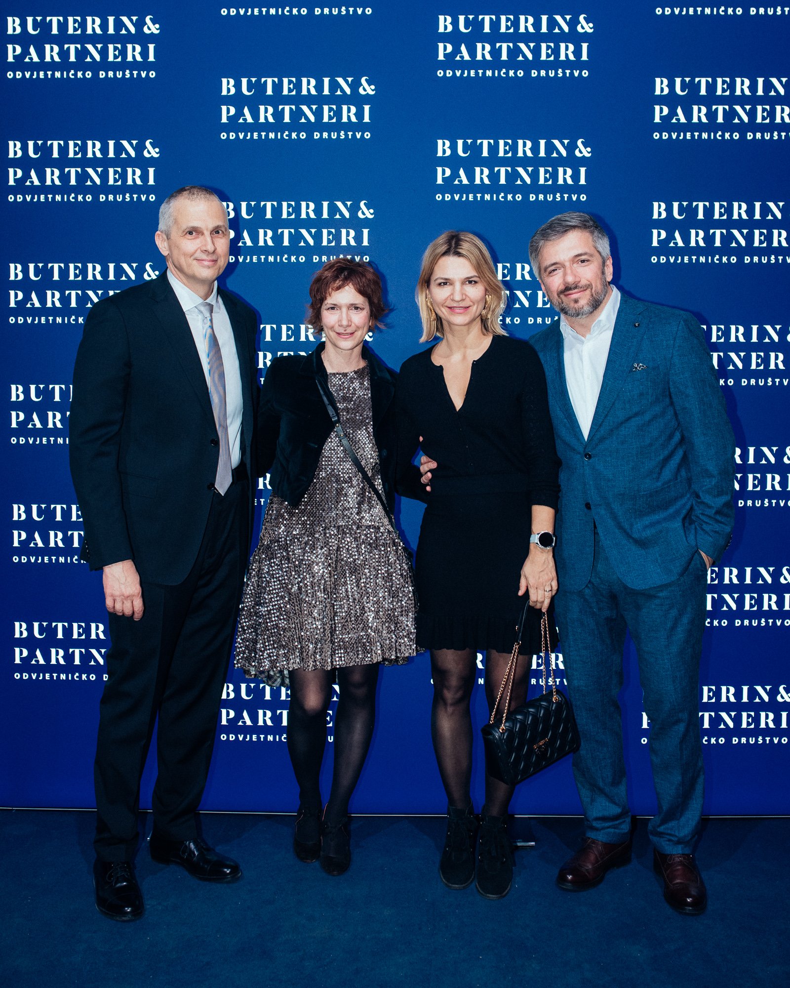 Four people standing together in front of a blue backdrop with white text, dressed in formal attire, at a social or professional event.