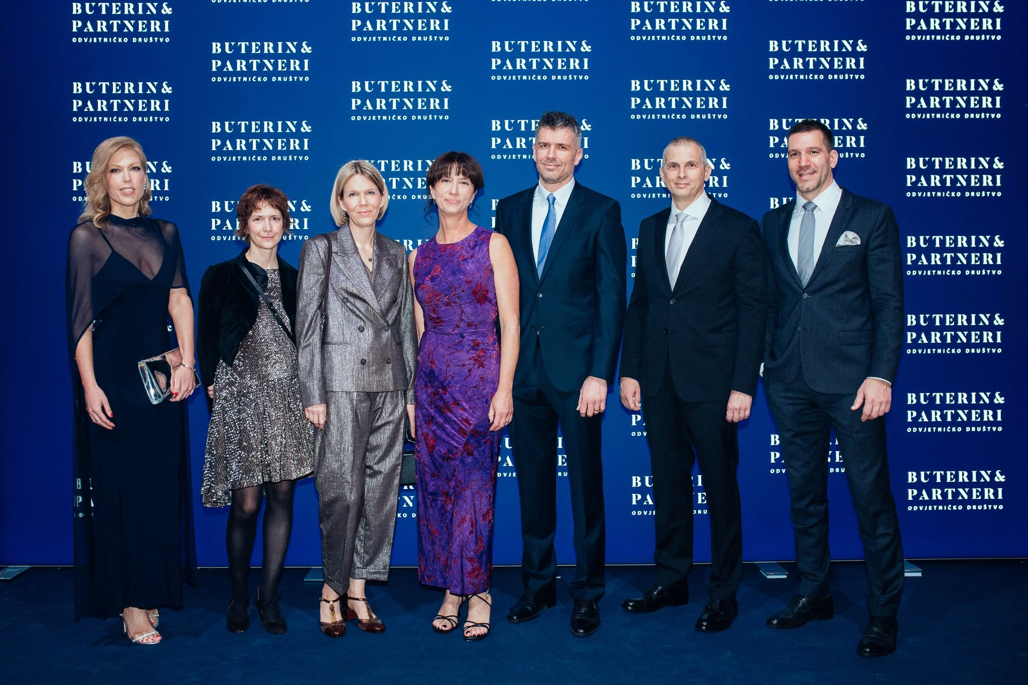 Eight people standing together in front of a blue backdrop with white text, dressed in formal attire, at a professional or corporate event.