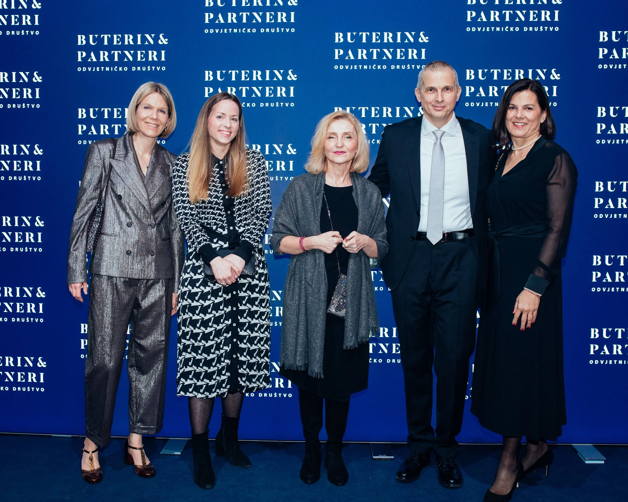 Group of six people standing in front of a blue backdrop with the logo and text "Buterin & Partneri" repeated in white, all dressed in formal attire, smiling for a photo at a professional event.