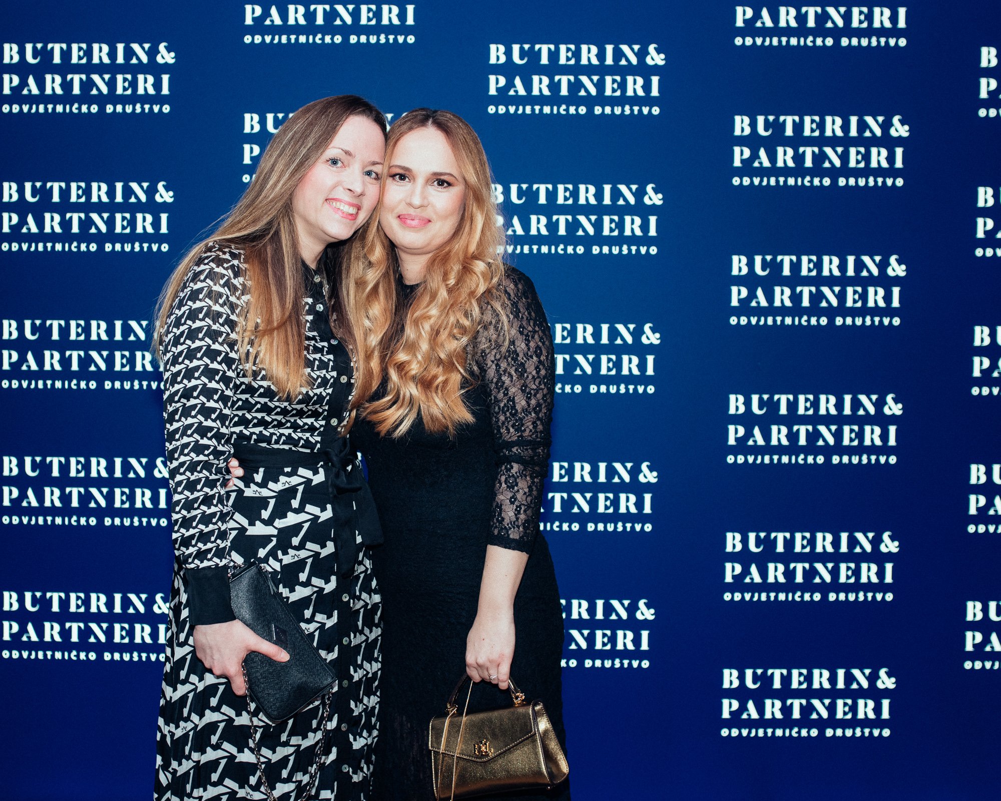 Two women standing together at an event, posing in front of a blue backdrop with white text that reads 'BUTERIN & PARTNERI ODVJETNICKO DRUŠTVO.' One woman has long blonde hair and is wearing a black and white patterned dress, holding a black clutch. 