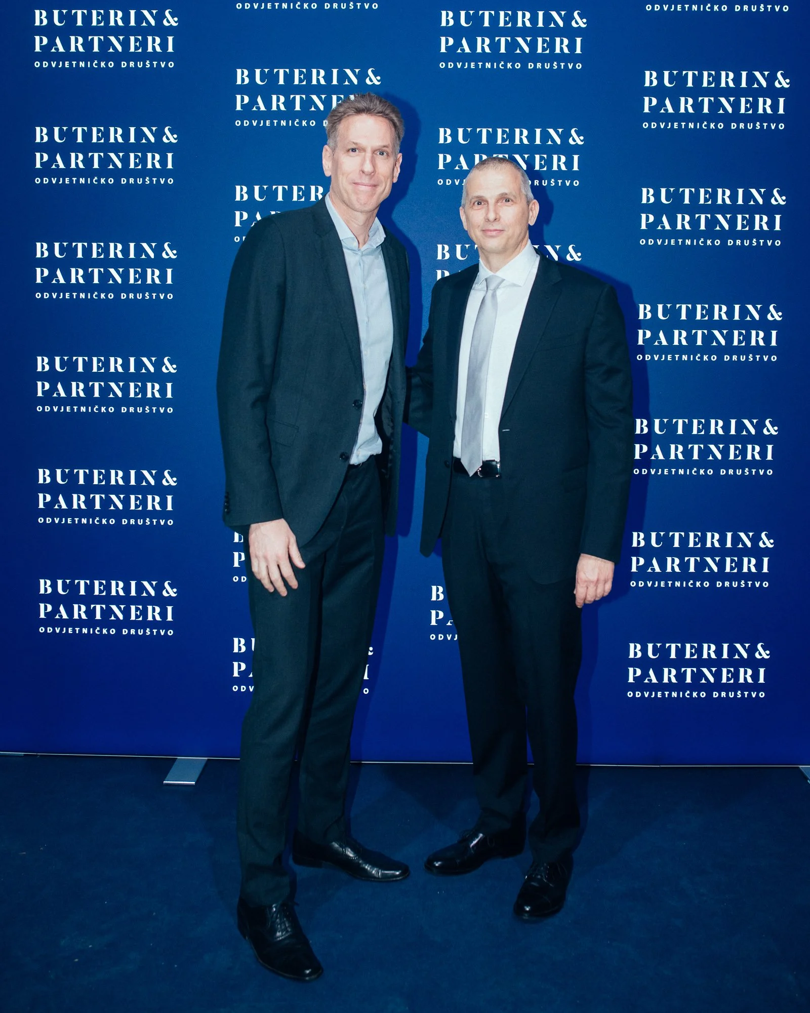 Two men in suits posing at a professional event with a blue backdrop that says 'Buterin & Partneri' repeatedly.