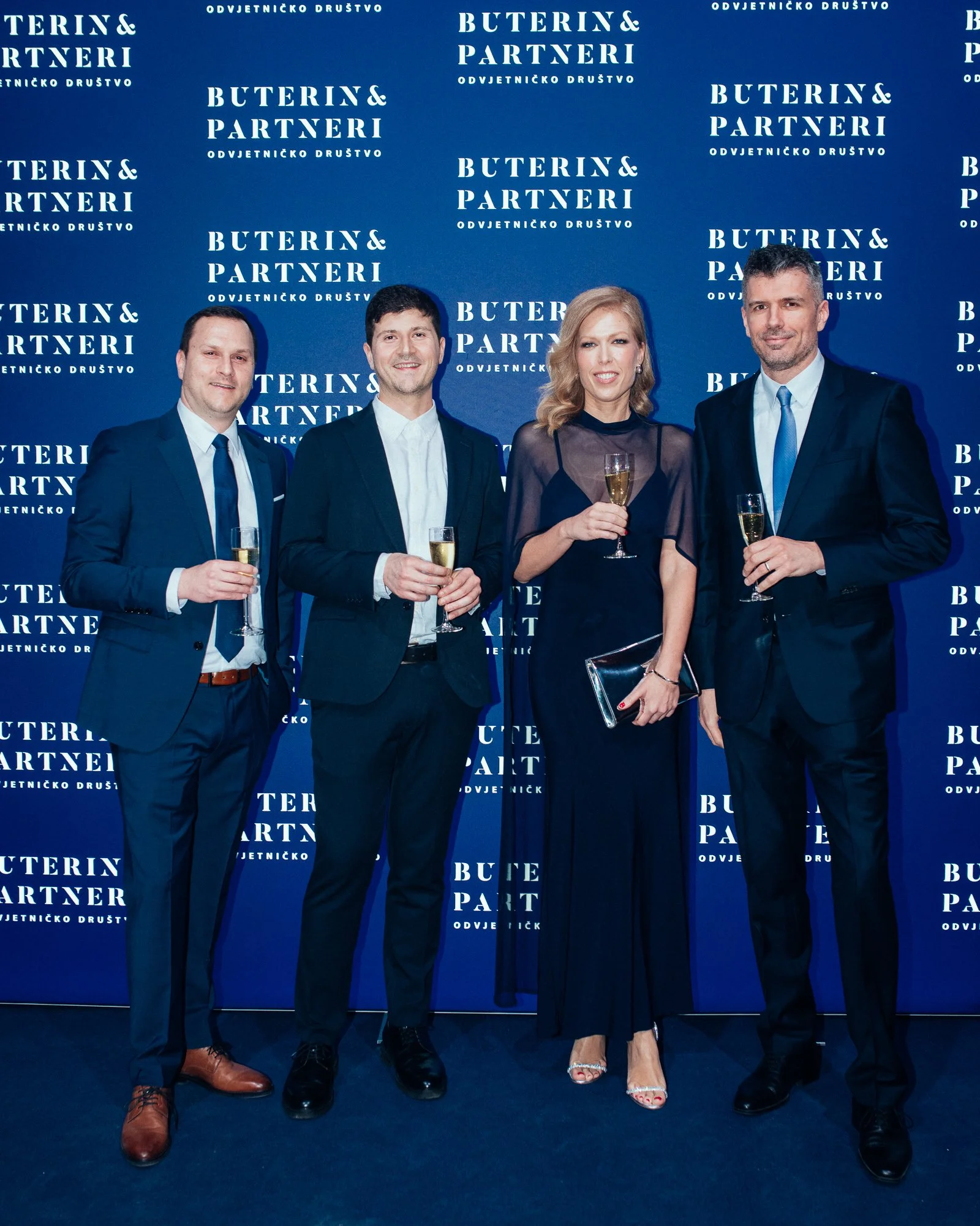 Four people in formal attire standing in front of a blue backdrop with white text that reads 'BUTERIN & PARTNERI ODVJETNIČKO DRUŠTVO'. They are holding glasses of champagne, smiling, and posing for the photo.
