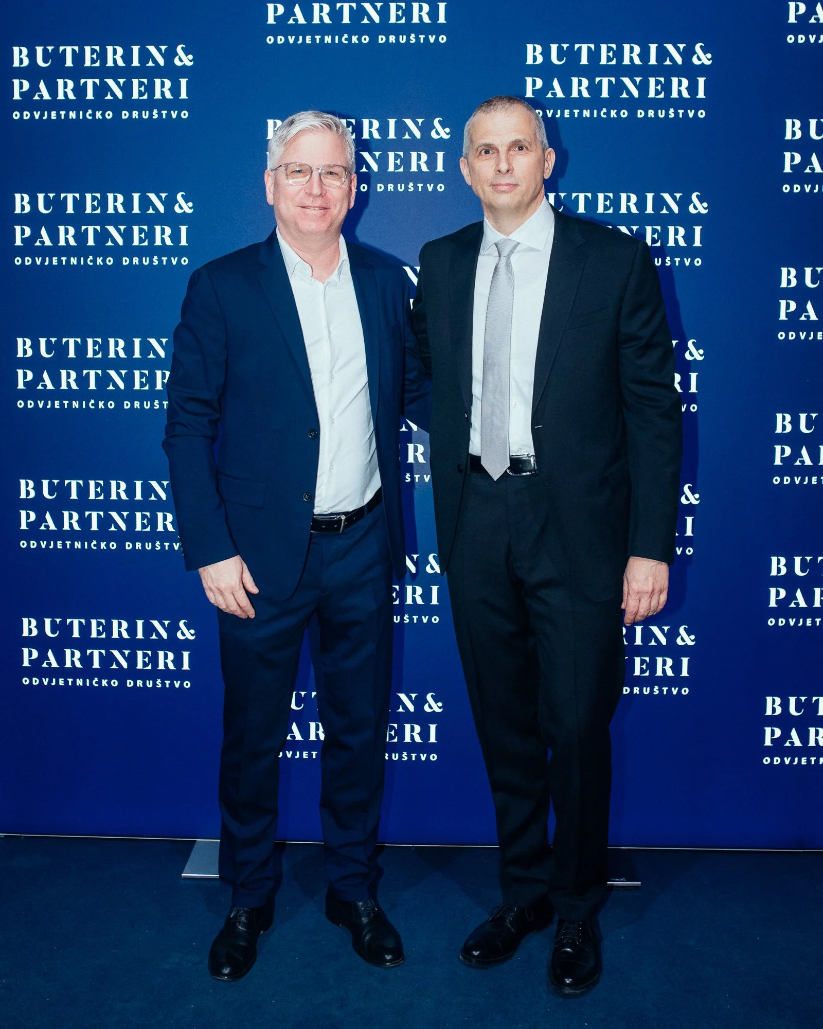 Two men in formal suits standing together in front of a blue backdrop with the words 'Buterin & Partneri' repeated, smiling at the camera.
