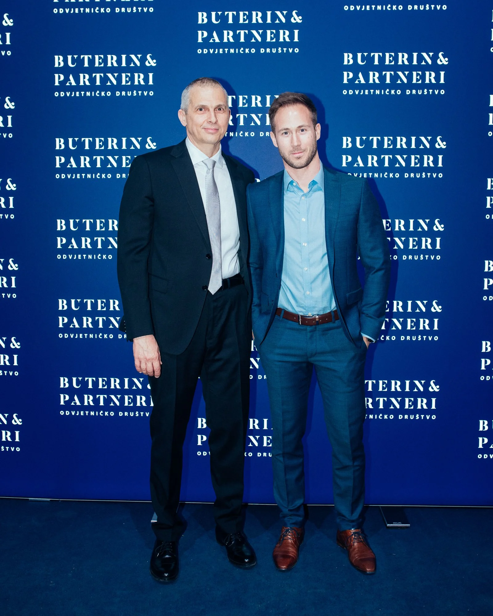 Two men in formal suits standing in front of a blue backdrop with white text that reads "BUTERIN & PARTNERI" and "ODVJETNIČKO DRUŠTVO." They are posing for a photo at what appears to be a professional or corporate event.