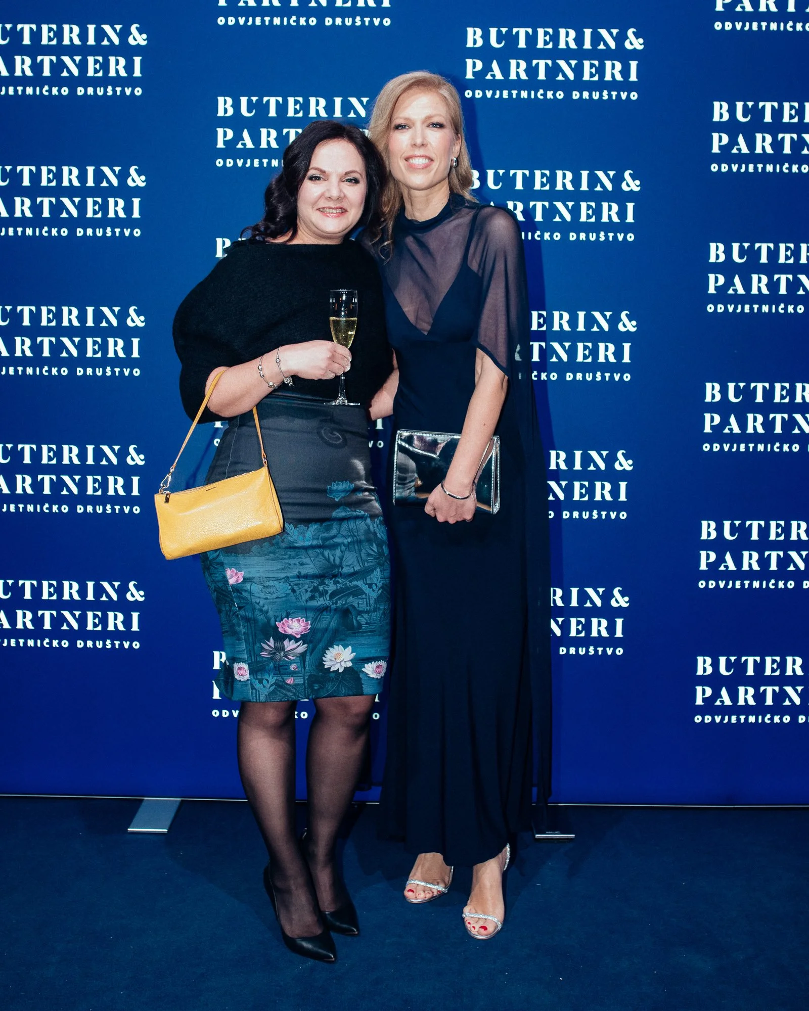 Two women dressed in evening attire posing together at an event, with a blue backdrop that reads "Buterin & Partneri" repeatedly. One woman is holding a glass of champagne, and both are smiling.