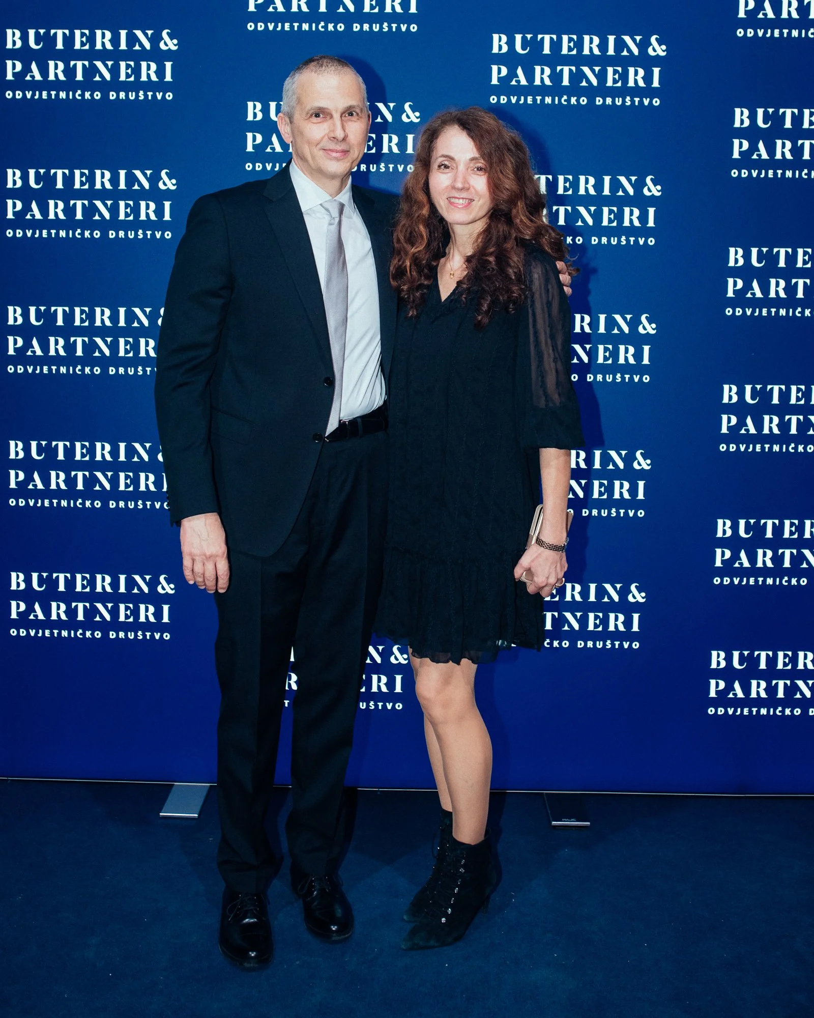 A man in a dark suit and a woman in a black dress are standing together in front of a blue backdrop with white text that reads 'Buterin & Partneri'. They are smiling and posing for the photo at an event.