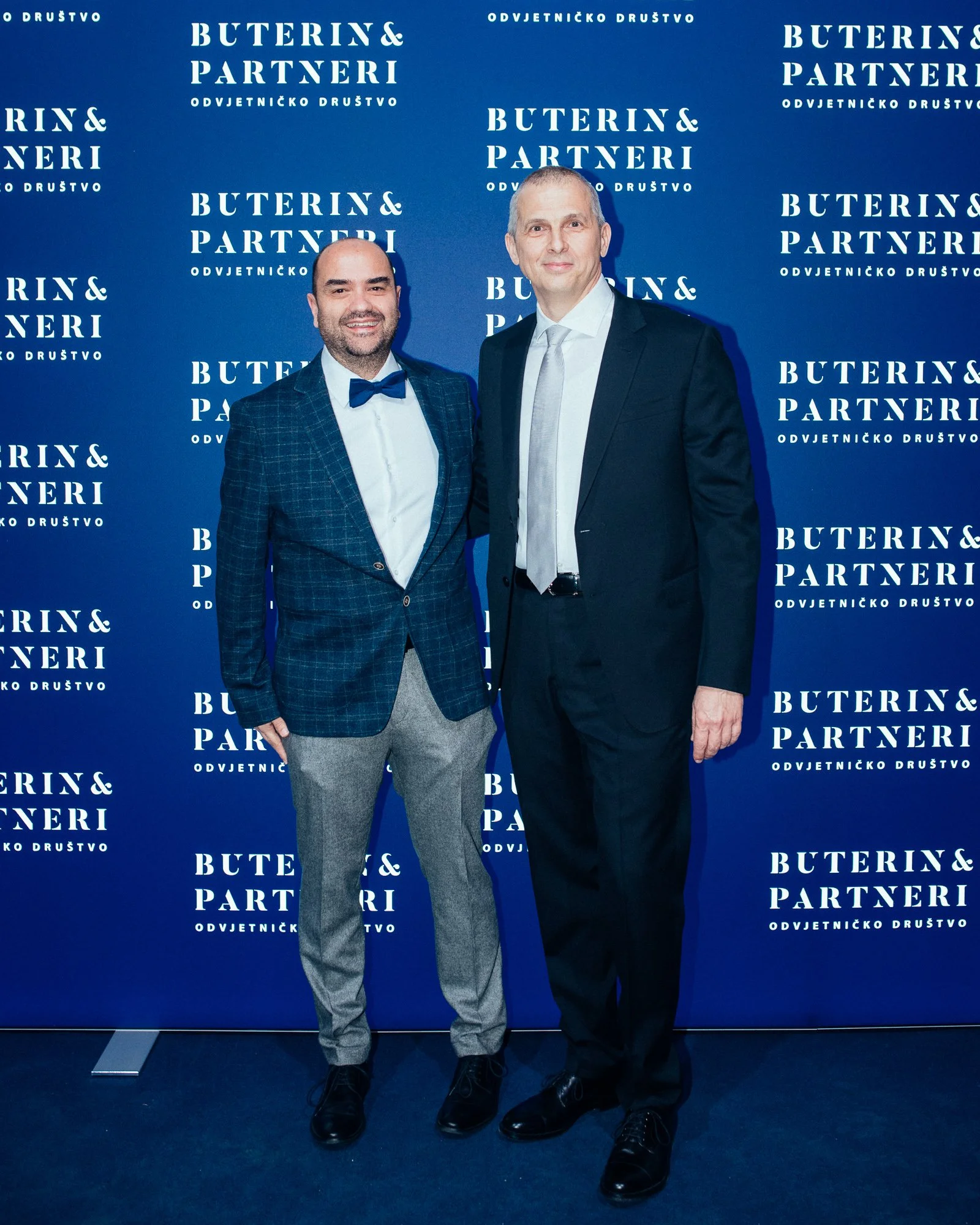 Two men in formal attire standing in front of a blue backdrop with white text that reads "Buterin & Partneri" and "Odvjetničko Društvo." One man is wearing a checked blazer, bow tie, and light grey pants, while the other is in a black suit with a whi