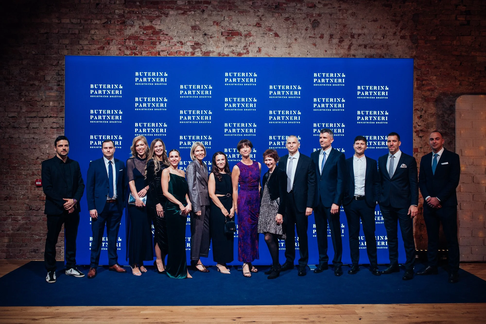 Group of fifteen people dressed in formal attire standing on a dark blue carpet in front of a blue backdrop with white text that reads "Buterin & Partneri" and other words, in a brick-walled room.