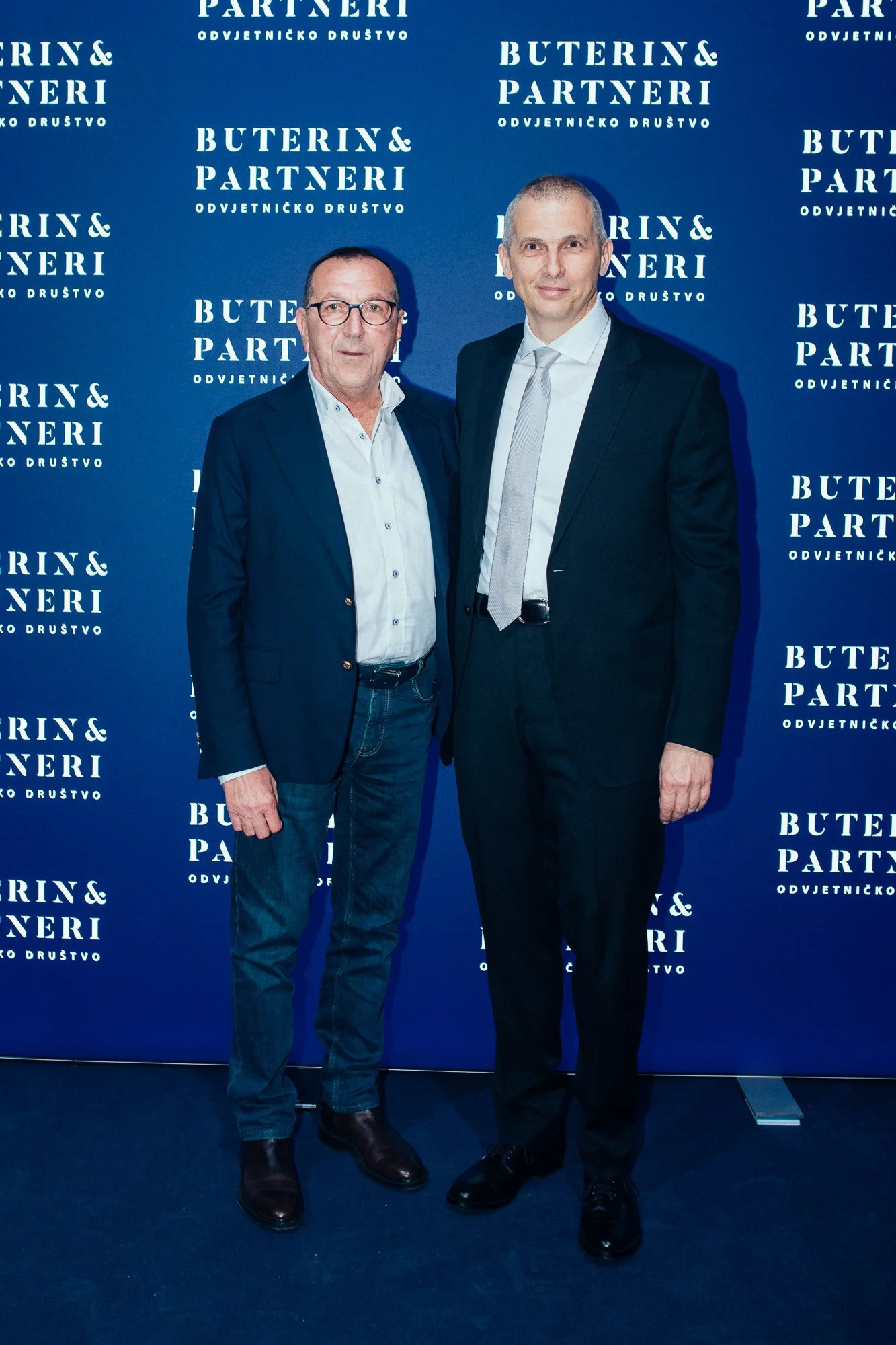 Two men standing side by side in front of a blue backdrop with white text that reads 'Buterin & Partneri'. The man on the left wears glasses, a dark blazer, light shirt, jeans, and black shoes. The man on the right is dressed in a dark suit, white sh