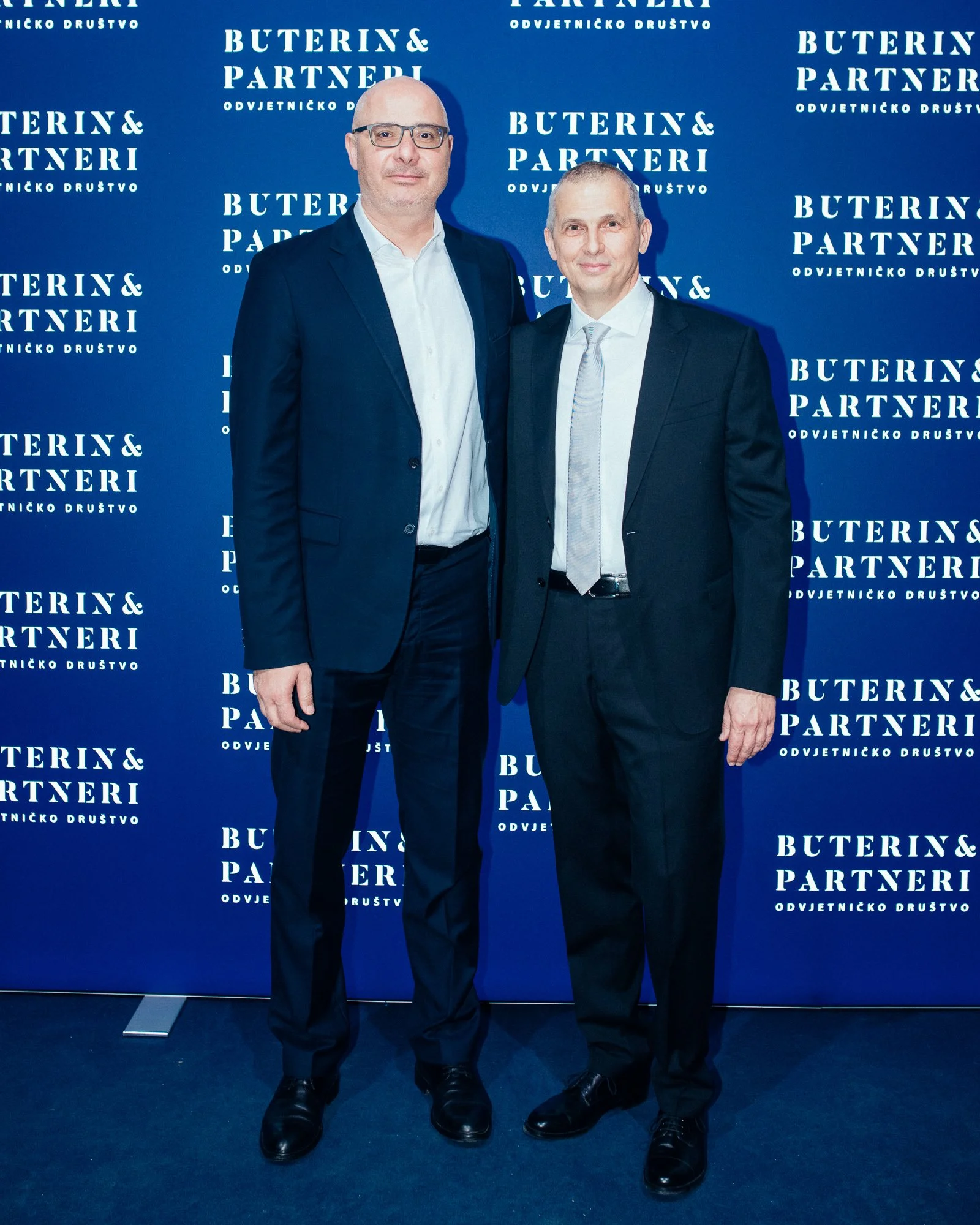 Two men in suits standing in front of a blue backdrop with white text that reads 'Buterin & Partneri Odvjetničko Društvo', smiling at the camera.
