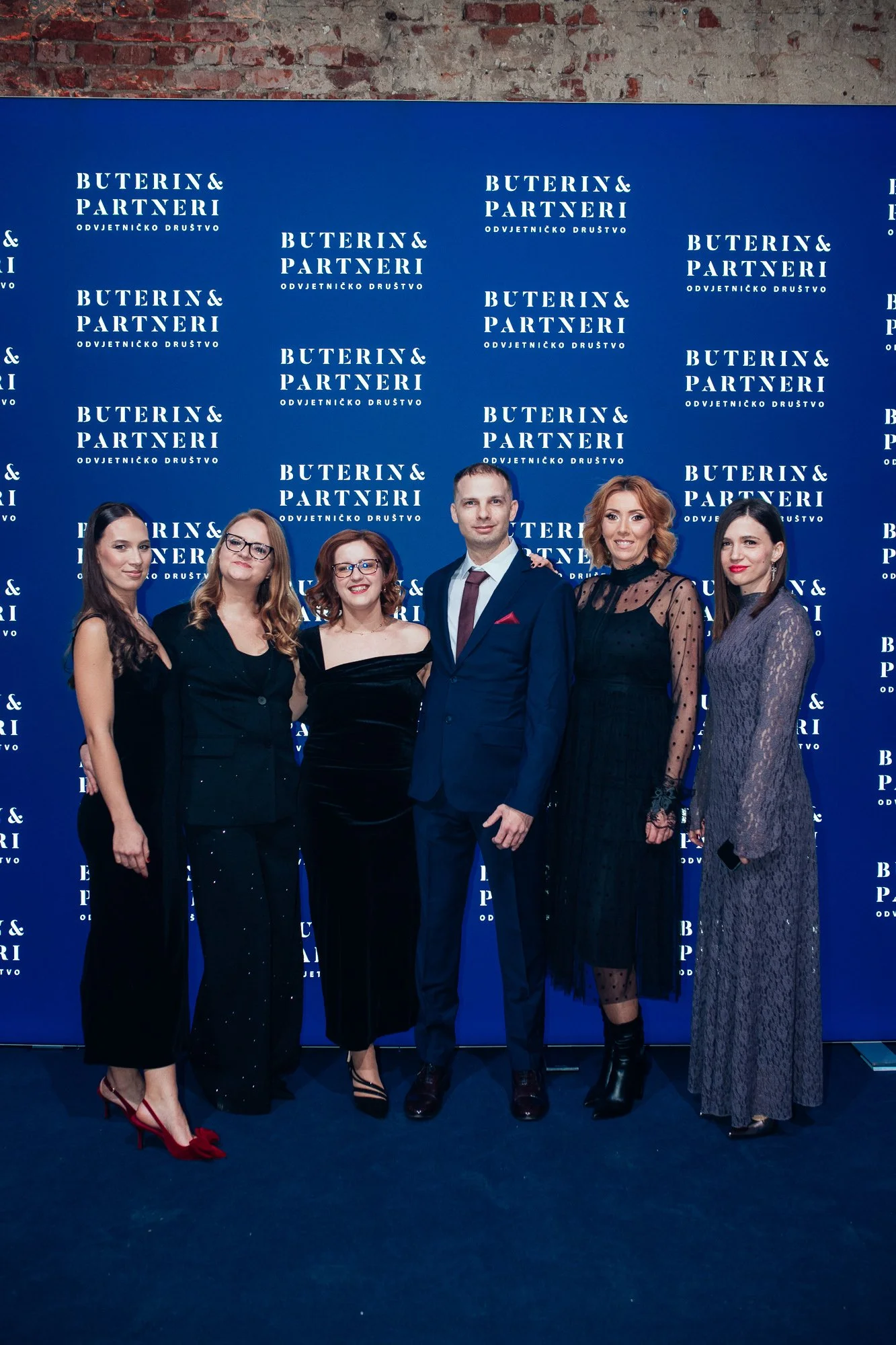 Group of six people dressed in formal attire standing in front of a blue backdrop with the repeated logo and text 'BUTERIN & PARTNERI ODVJETNIČKO DRUŠTVO'
