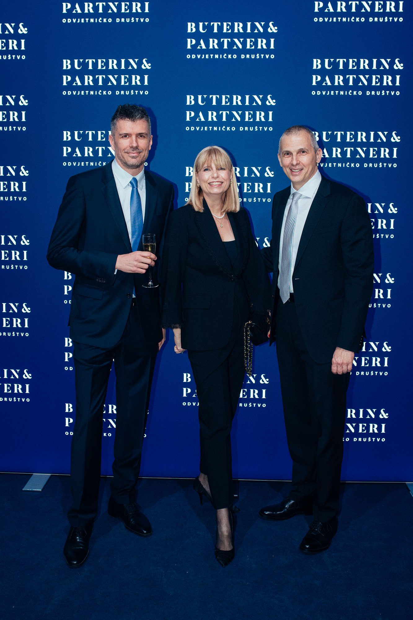 Three people in formal attire standing in front of a blue backdrop with white text that reads 'Partners Bond & Partners, Odvjetničko društvo.' The man on the left is holding a glass of champagne. All three are smiling and posing for the photo.