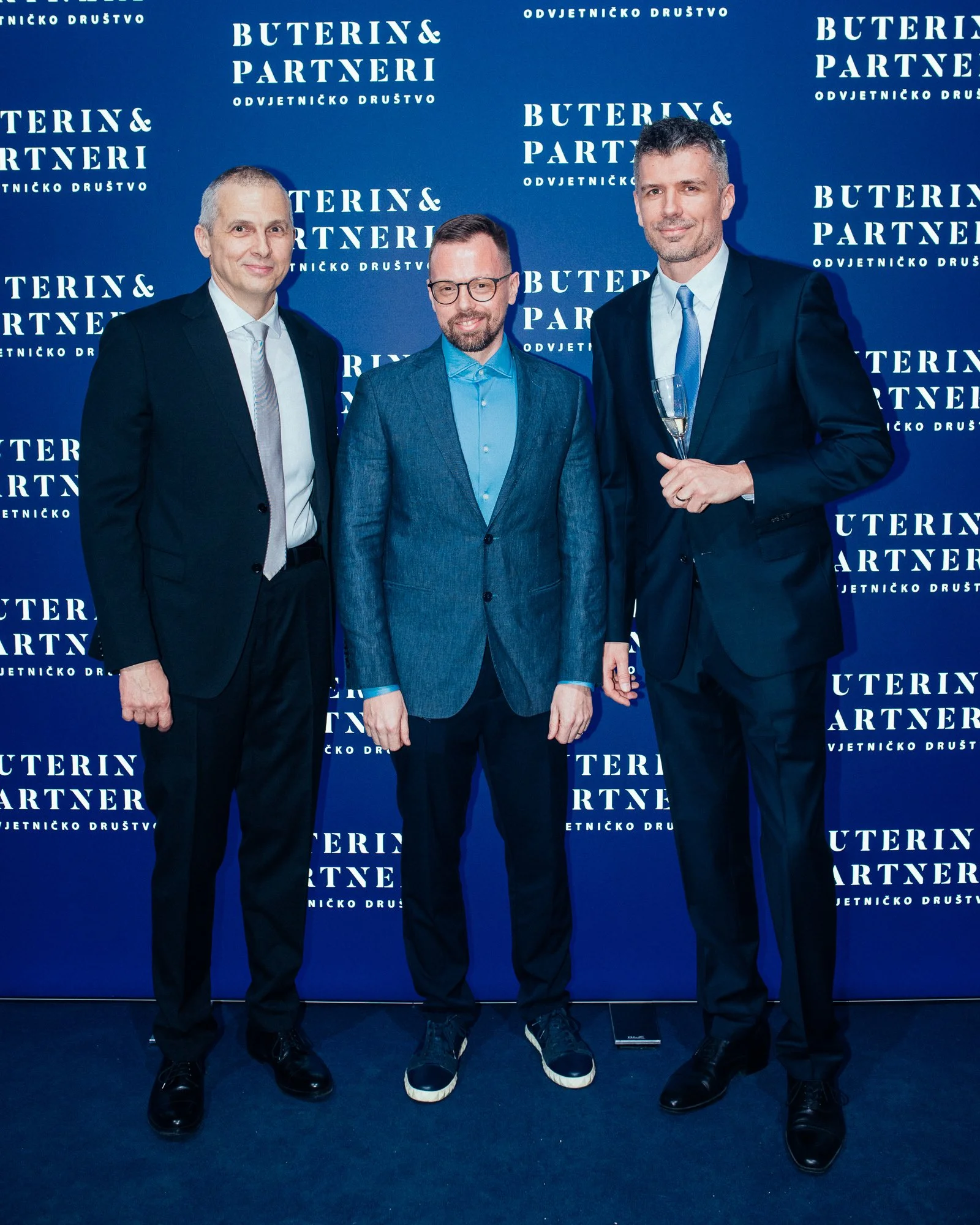 Three men in suits standing in front of a blue backdrop with white text that reads 'Buterin & Partneri' and 'Odvjetničko Društvo.' One man is holding a glass of champagne.