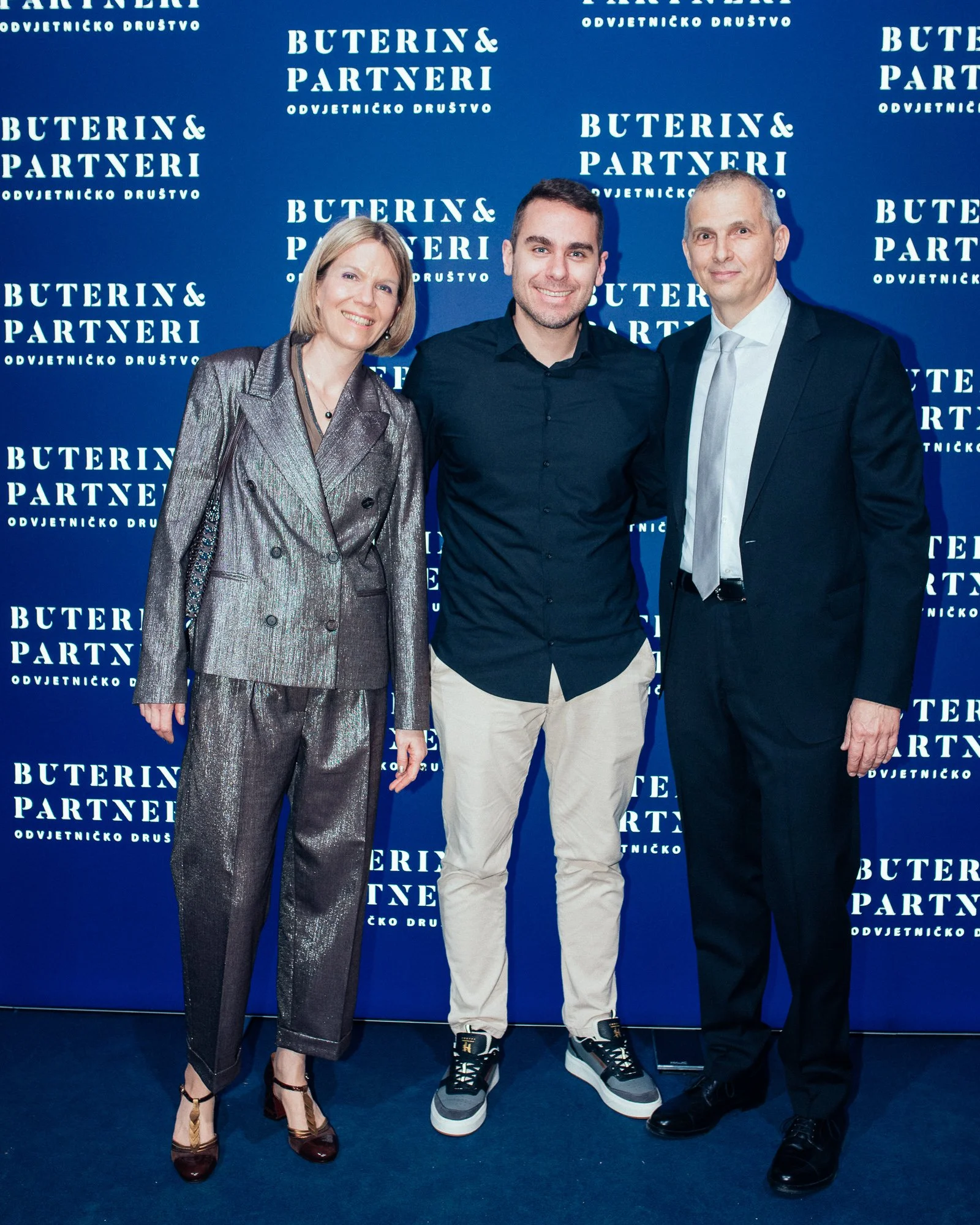 Three people standing in front of a blue backdrop with white text that reads 'Buterin & Partneri'. The woman on the left is wearing a shiny silver pantsuit and high heels, the man in the middle is dressed in a black shirt and beige pants, and the man