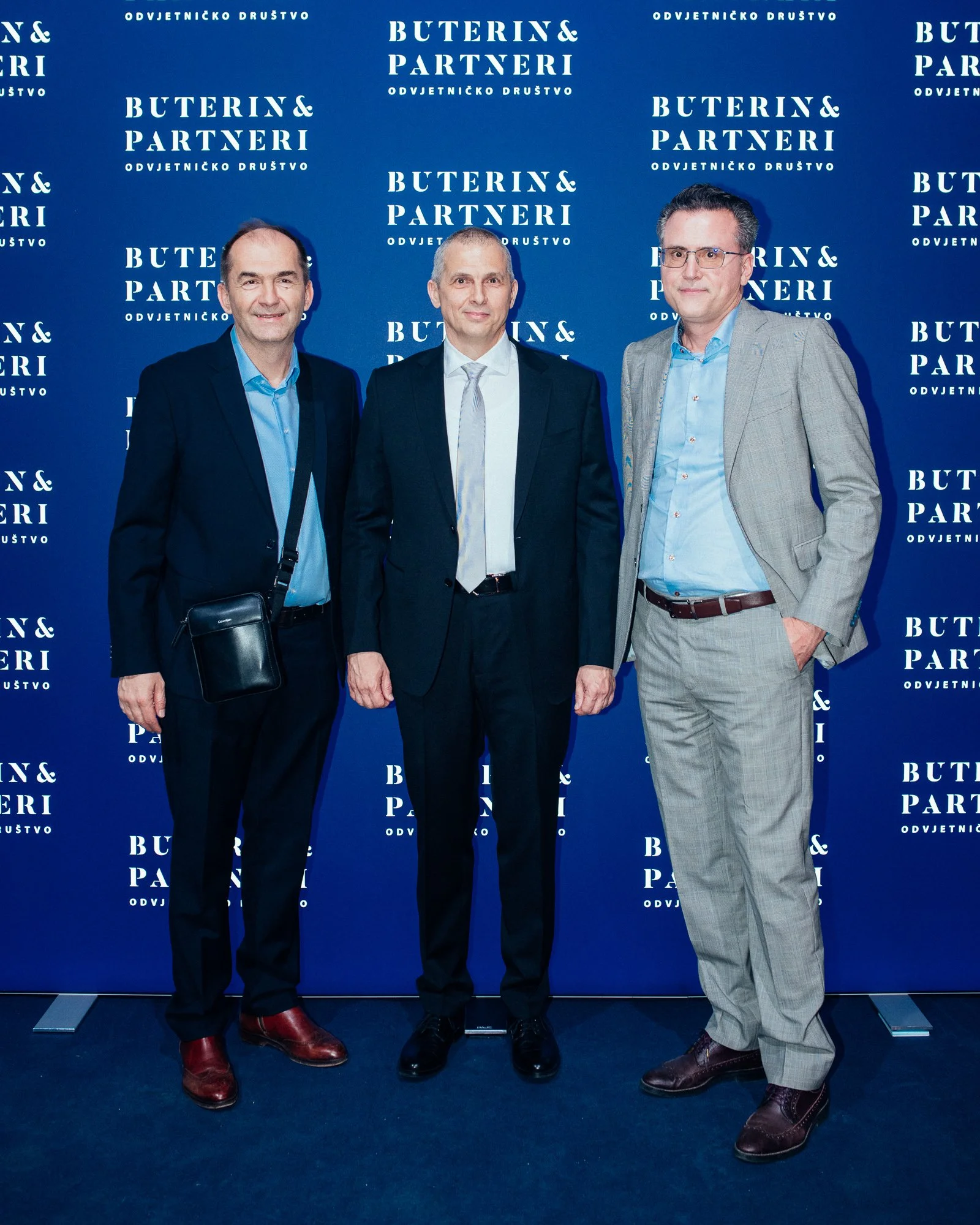 Three men in formal attire standing in front of a blue backdrop with the text 'BUTERIN & PARTNERI' repeated multiple times, posing for a photo.