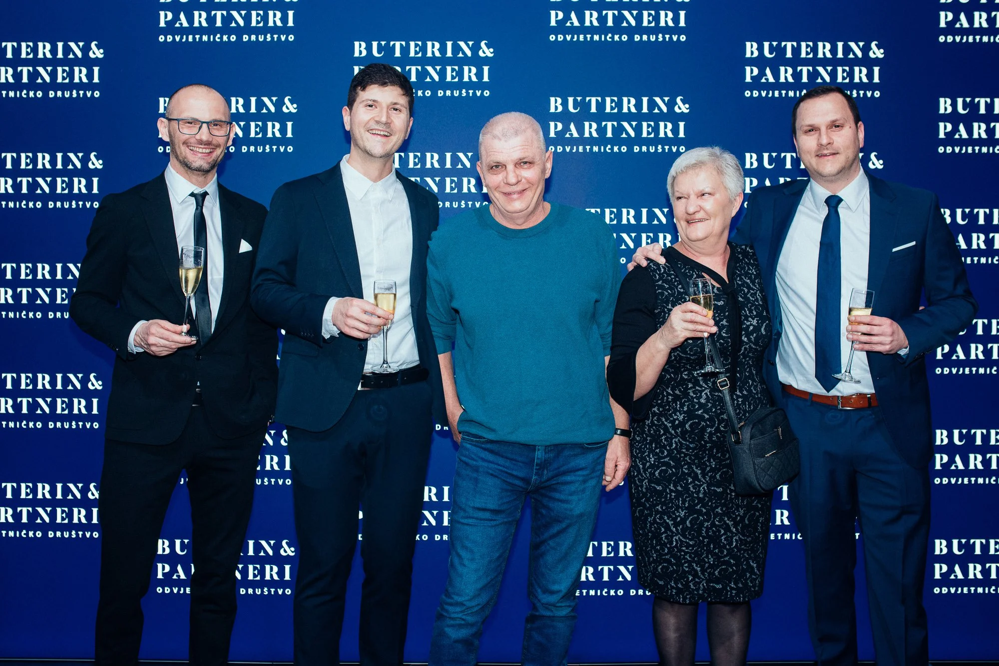 A group of six people at an event, standing in front of a blue backdrop with white text. They are dressed in formal and semi-formal attire, holding glasses of champagne, smiling, and posing for the photo.