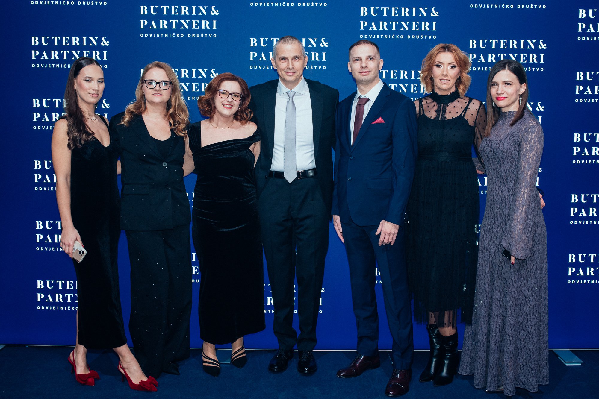Group of seven people dressed in formal attire, standing in front of a blue backdrop with the text "Buterin & Partneri".
