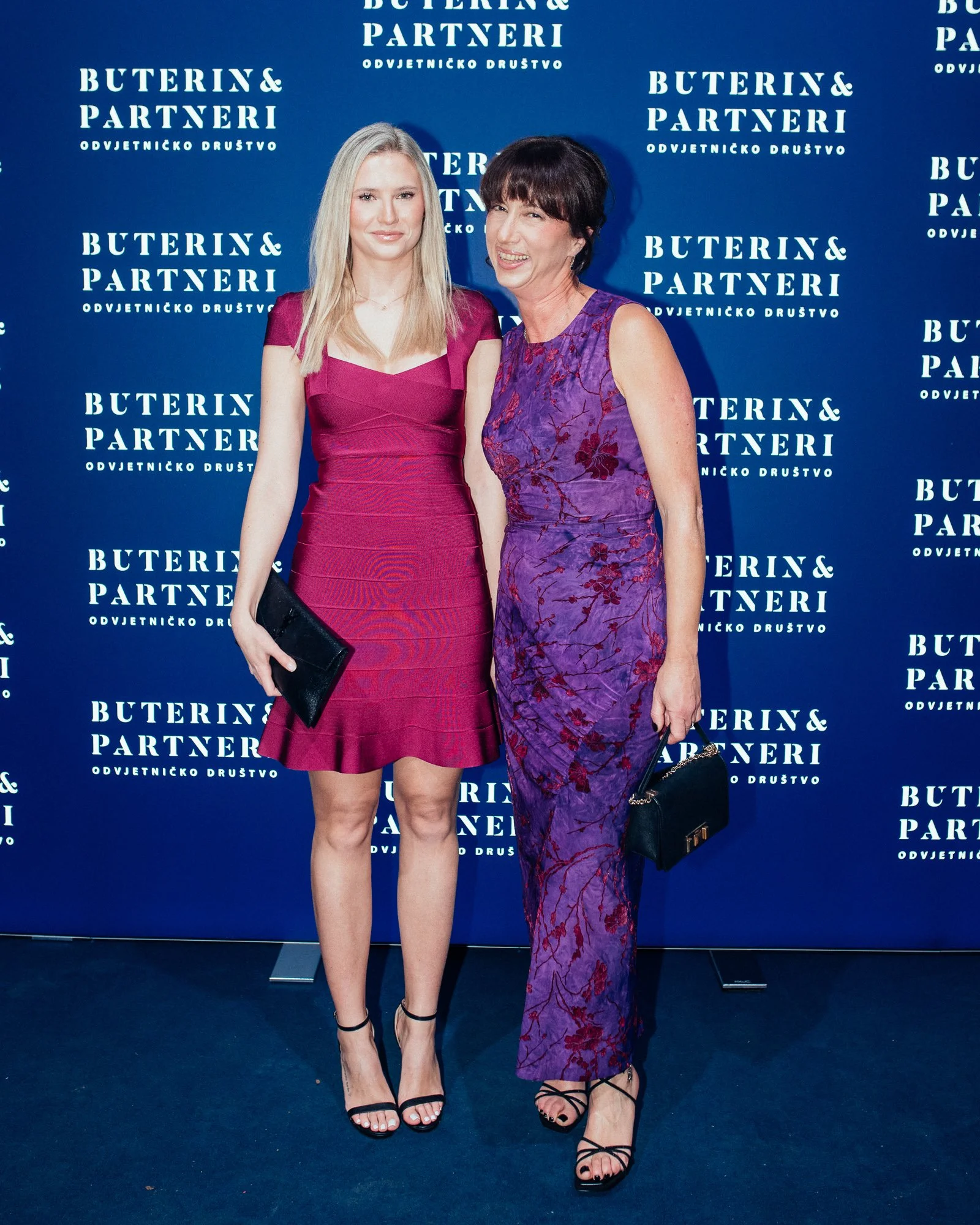 Two women standing together posing for a photo in front of a blue backdrop with the words 'BUTERIN & PARTNERI' repeated multiple times. One woman is wearing a fitted pink dress with black high-heeled sandals, holding a black clutch. The other woman i