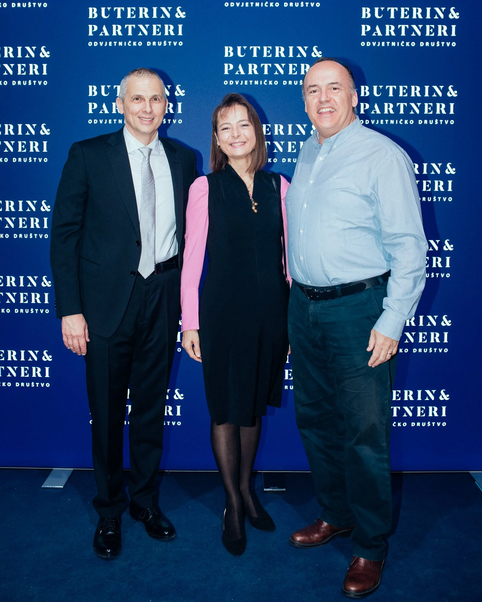 Three people standing in front of a blue backdrop with white text that reads "Buterin & Partneri, Odvjetničko društvo." The person on the left is a man in a dark suit and tie, the person in the middle is a woman in a black dress with pink sleeves, an