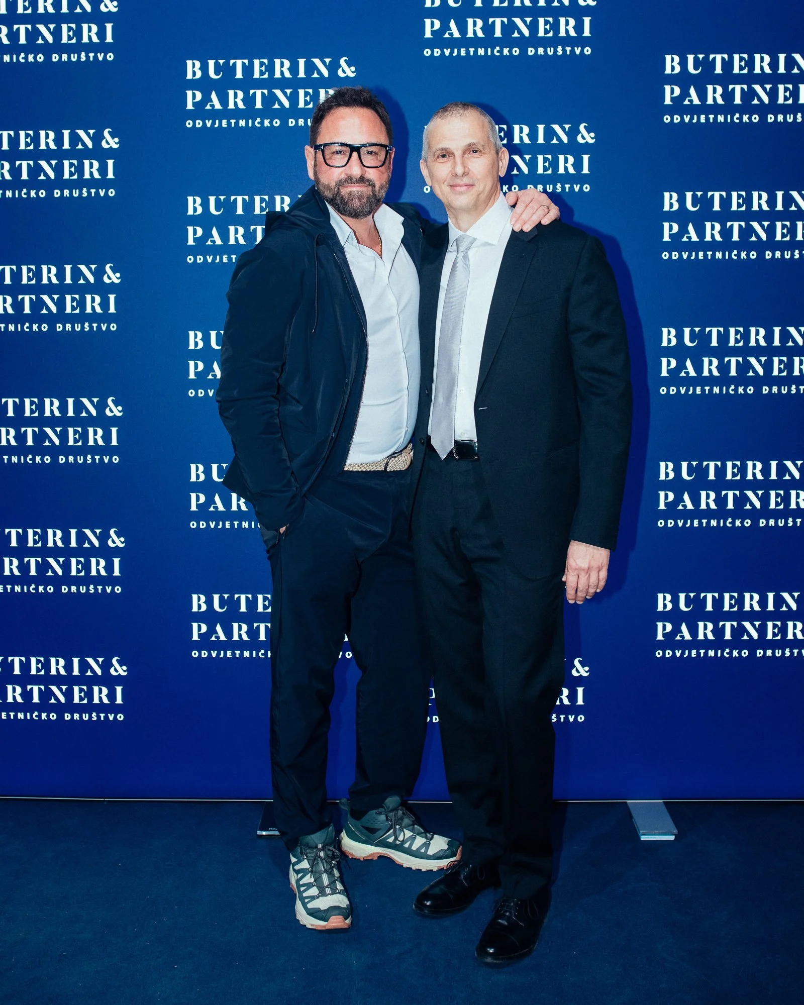 Two men standing together at a formal event with a blue background that reads "Buterin & Partneri" repeatedly. One man is wearing a black suit with a white shirt and black shoes; the other is dressed casually in a jacket, white shirt, and sneakers.
