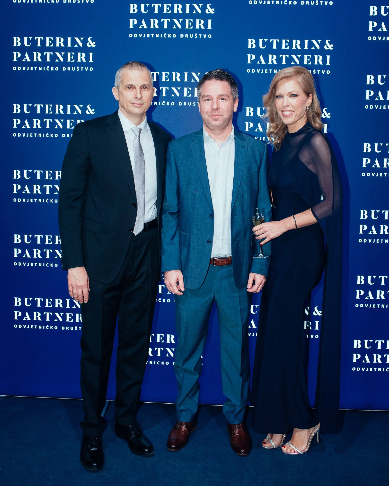 Three people in formal attire posing in front of a blue backdrop with white text that reads 'Buterin & Partneri' and 'Odvjetničko Društvo'. The person on the right is holding a glass.