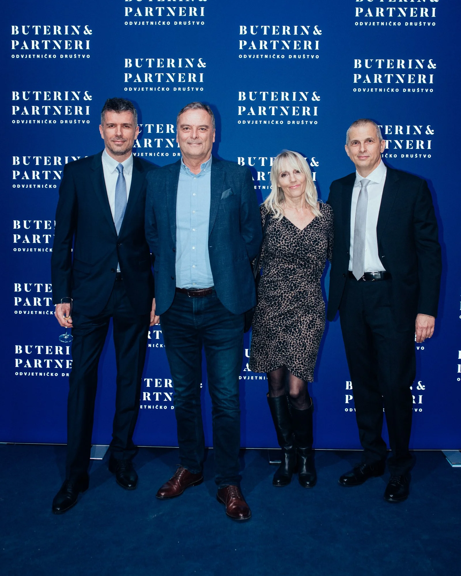 Four business professionals standing in front of a blue backdrop with white text that reads 'Buterin & Partneri Odvjetničko Društvo.' They are dressed in formal attire, with three men in suits and a woman in a leopard-print dress, all smiling at the 