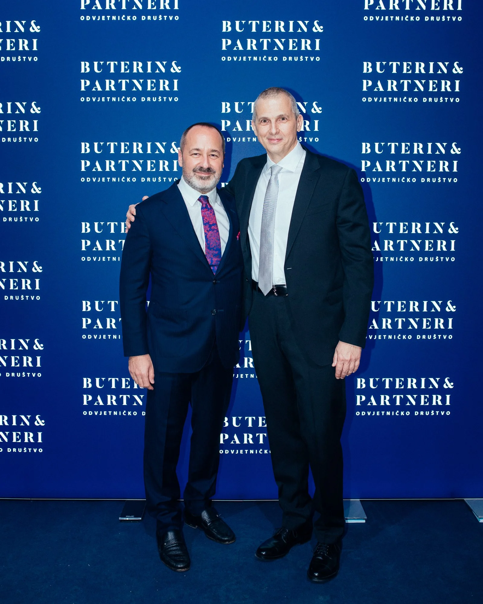 Two men in suits standing together in front of a blue backdrop with the text 'Buterin & Partneri' and 'Odvjetnicko Drustvo' repeating. One man has a beard and is wearing a dark blue suit with a pink and purple patterned tie. The other is taller, clea