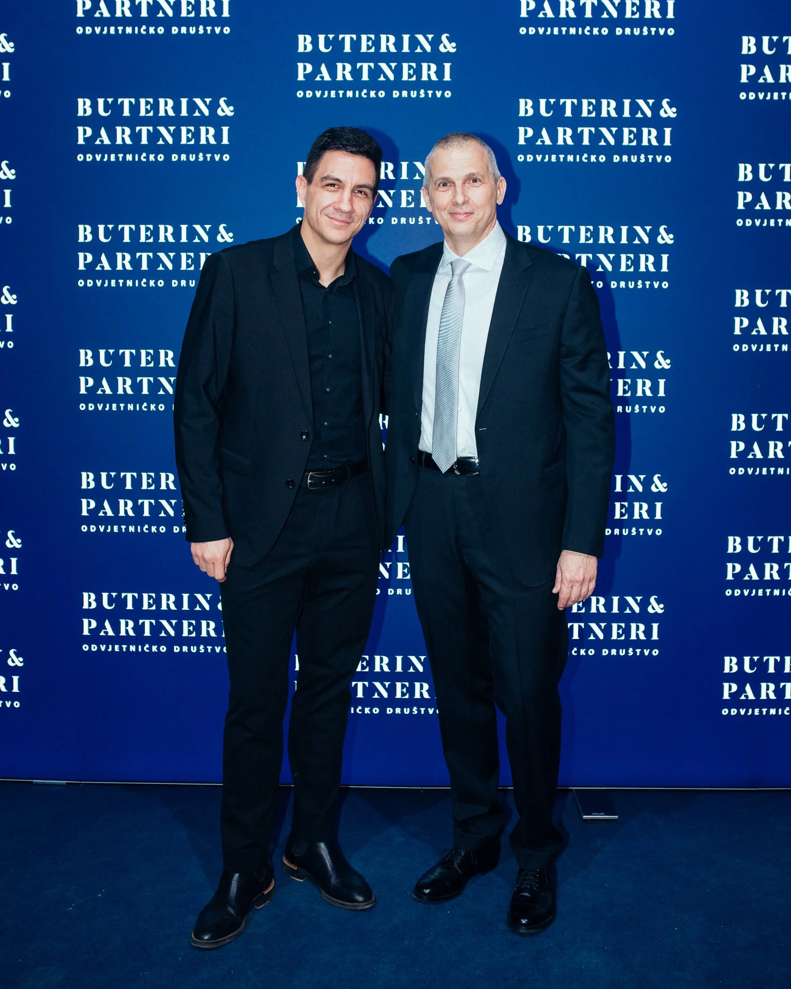 Two men in formal suits standing together at an event, posing for a photo in front of a blue backdrop with the text 'Buterin & Partneri' and other words written repeatedly.