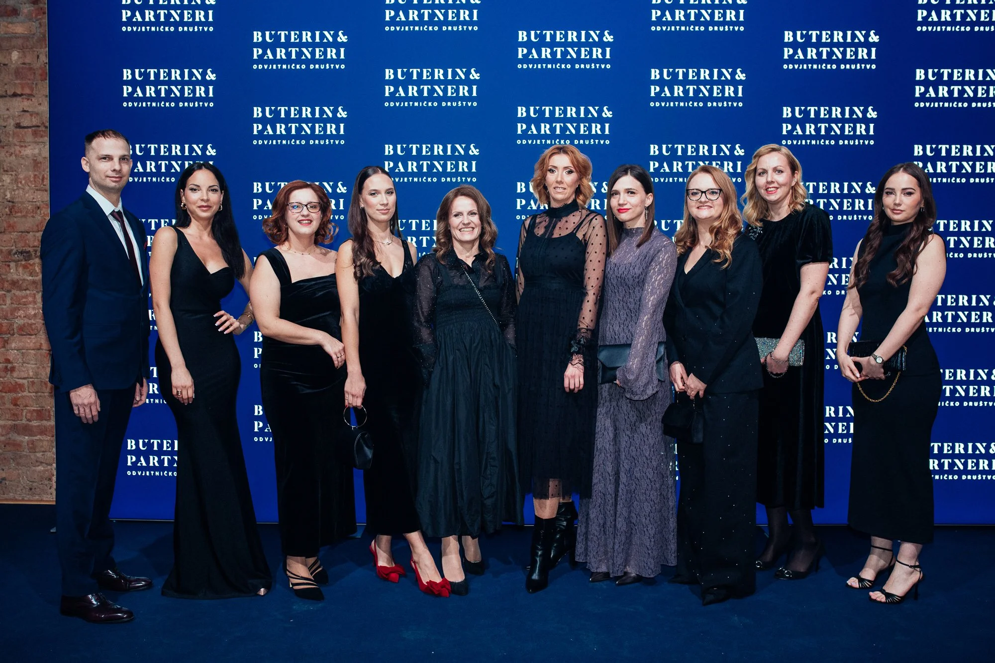 Group of eleven women and one man standing in front of a blue backdrop with white text, dressed in formal black attire, at an event or gathering.