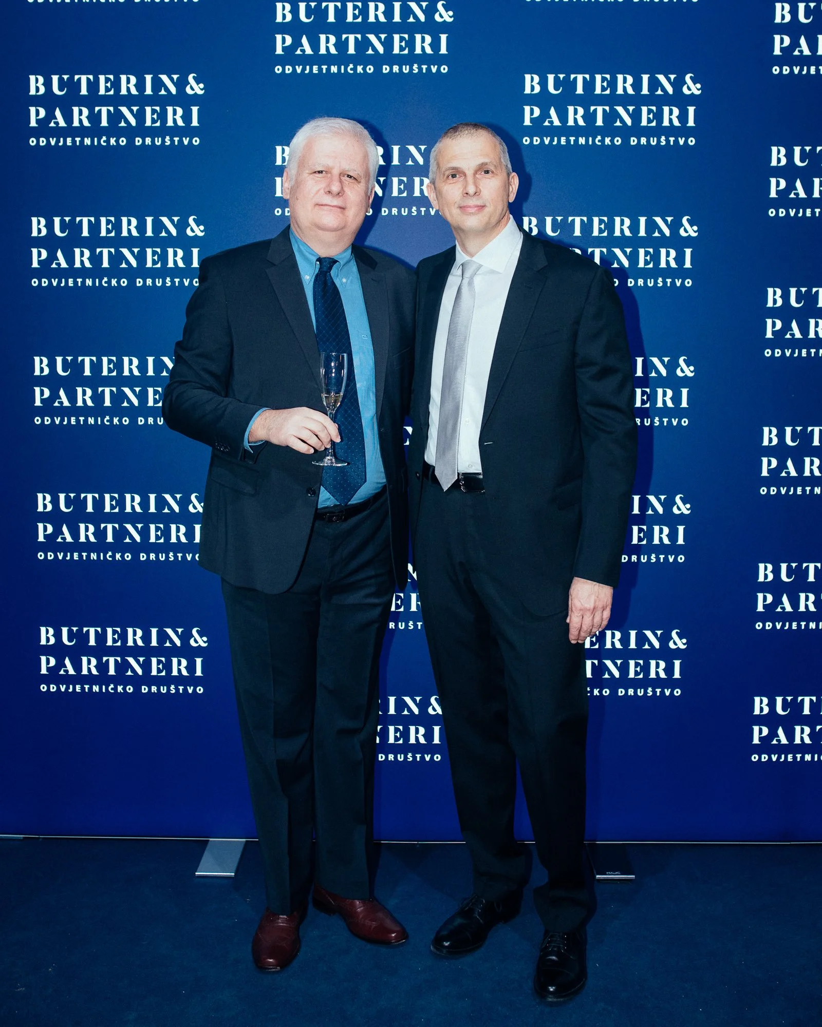 Two men in suits posing in front of a blue backdrop with white text that reads 'Buterin & Partneri Odvjetničko Društvo.' One man is holding a champagne flute.