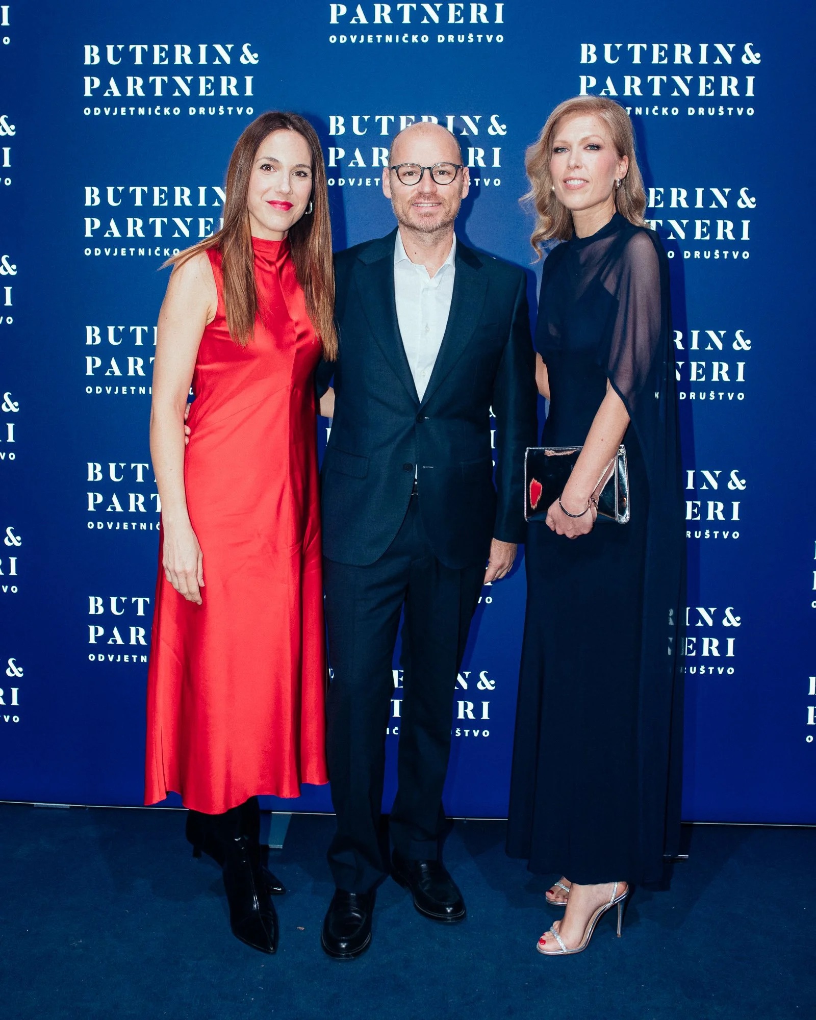 Three people standing in front of a blue backdrop with white text that reads 'BUTERIN & PARTNERI' and other Croatian words. The woman on the left is wearing a sleeveless red satin dress and black boots. The man in the middle is wearing a dark suit wi