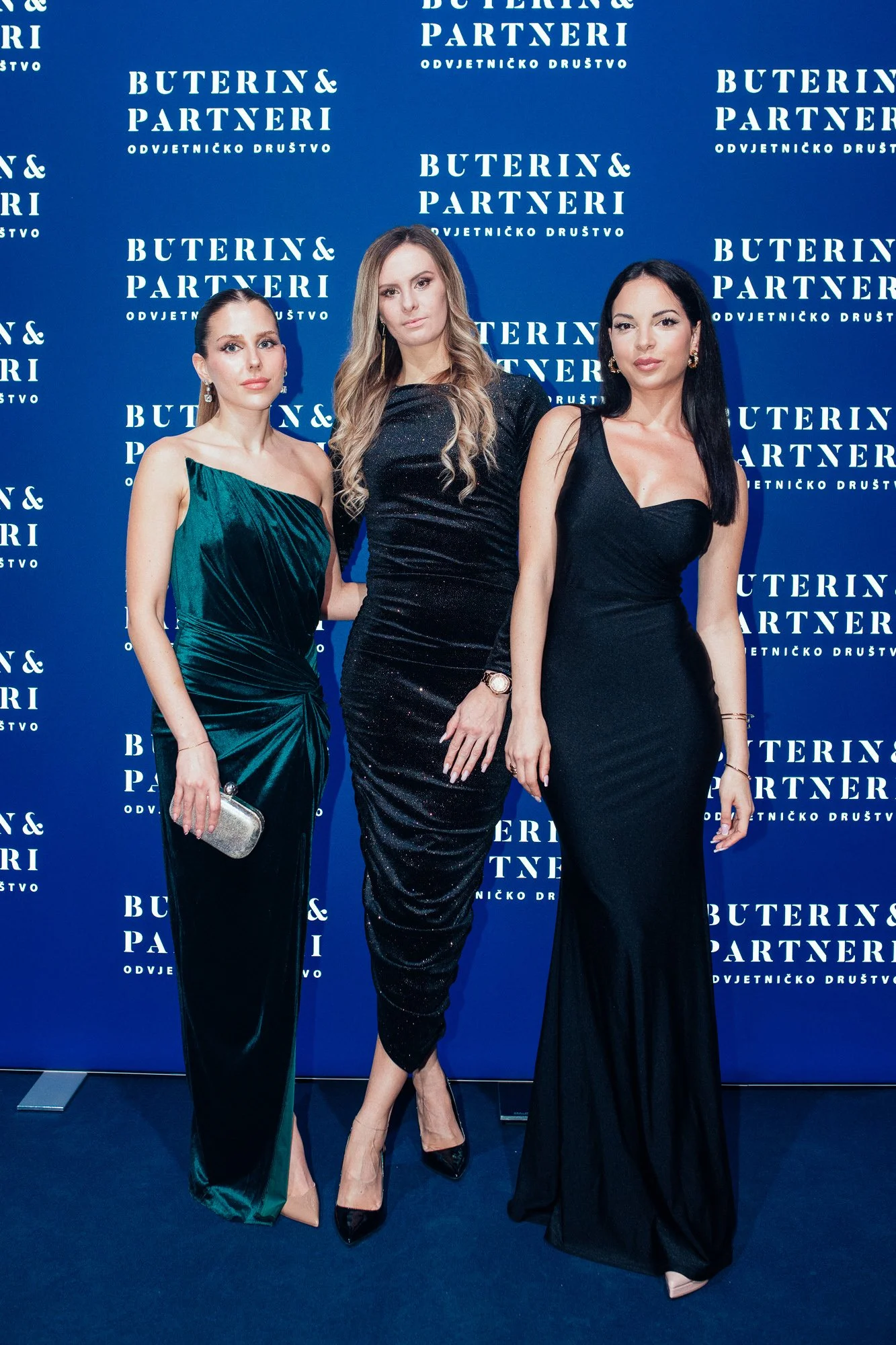 Three women in elegant evening dresses standing in front of a blue backdrop with white text that reads 'Buterin & Partneri Odvjetničko Društvo.'