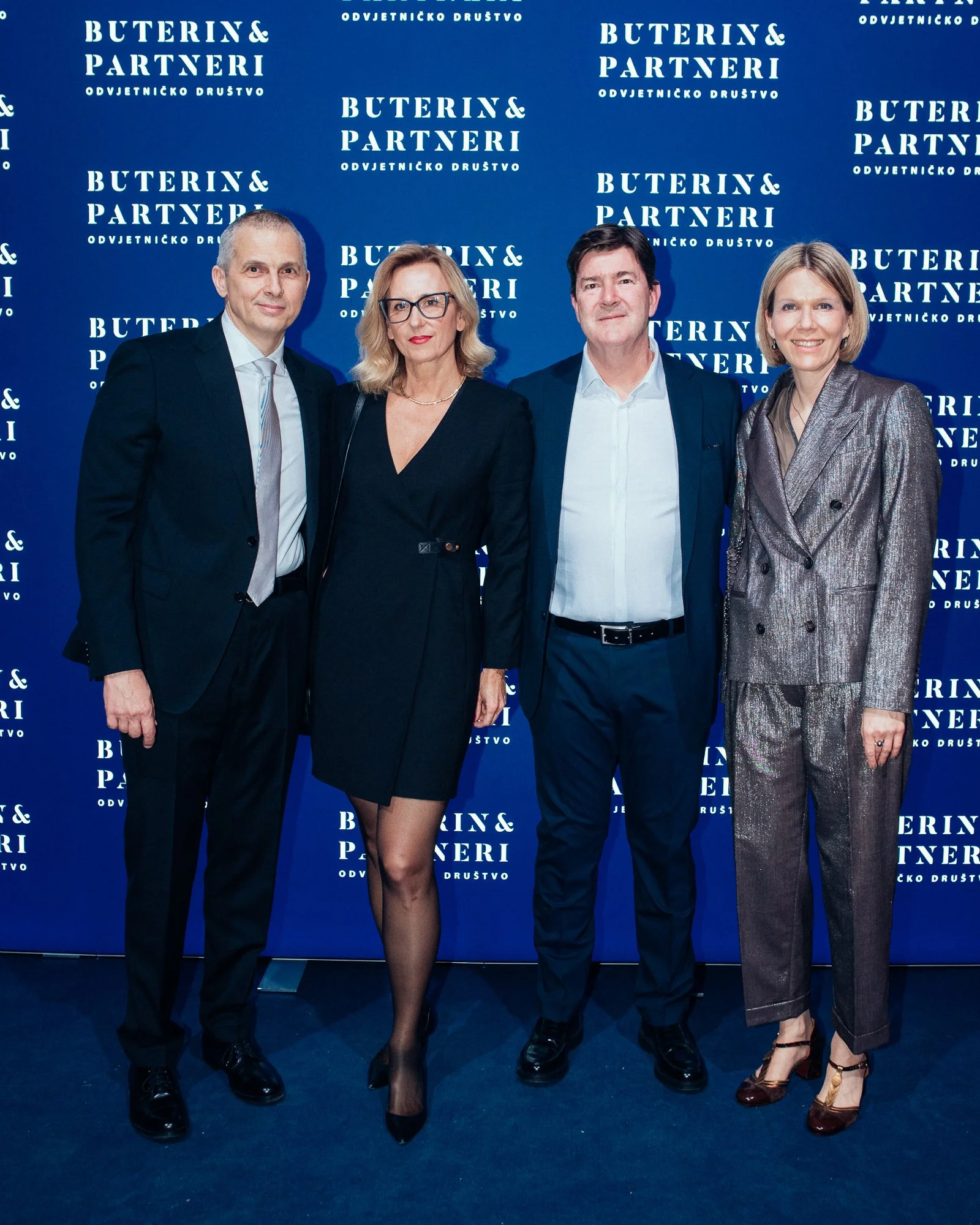 Four people standing in front of a blue backdrop with the text 'BUTERIN & PARTNERI.' They are dressed in formal attire, with the two women wearing dresses and the two men in suits.