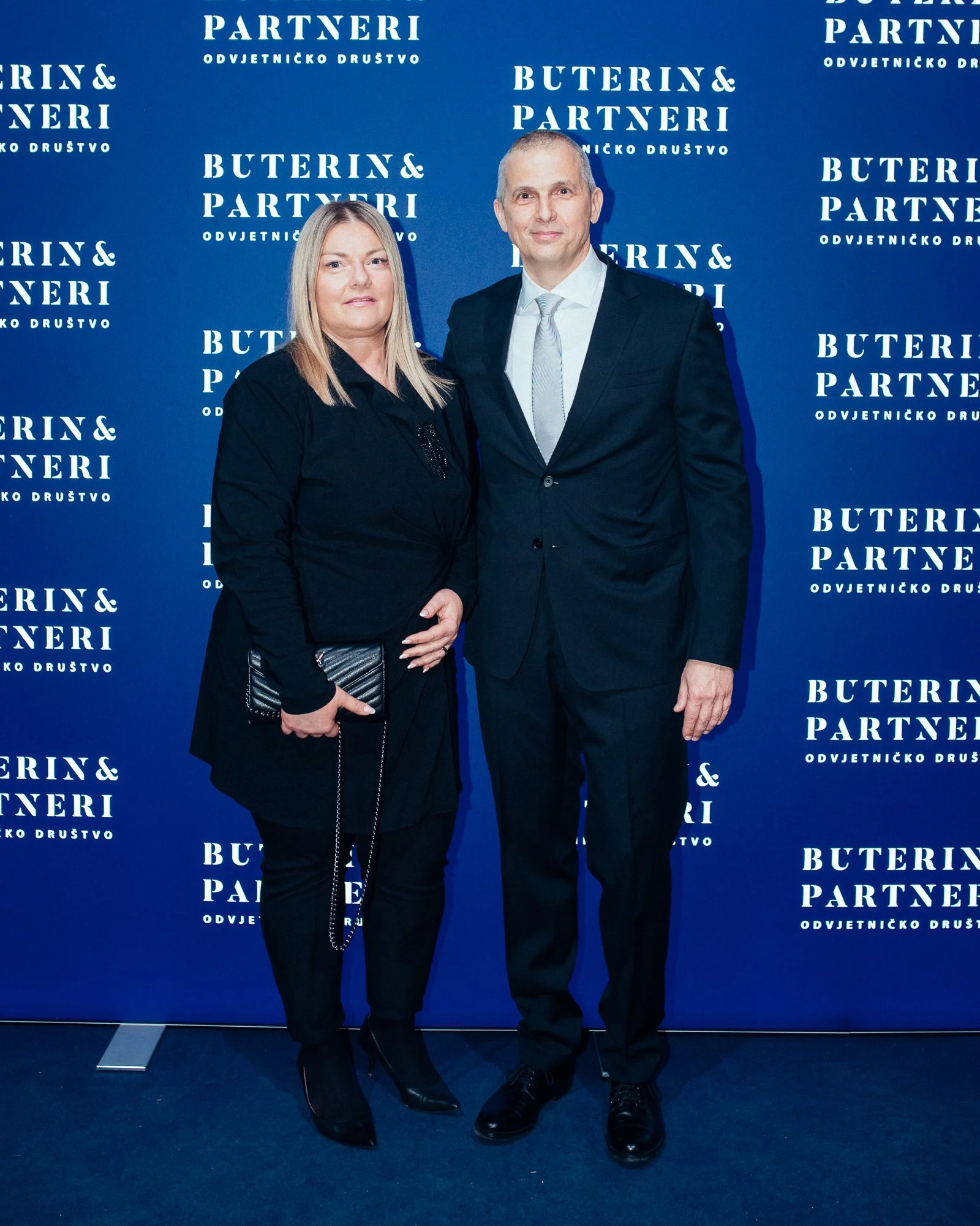 A man and woman standing together at a formal event with a blue backdrop featuring the words 'Buterin & Partneri, Odvjetničko Društvo' repeatedly printed on it. The woman is dressed in black holding a clutch, and the man is wearing a suit and tie.