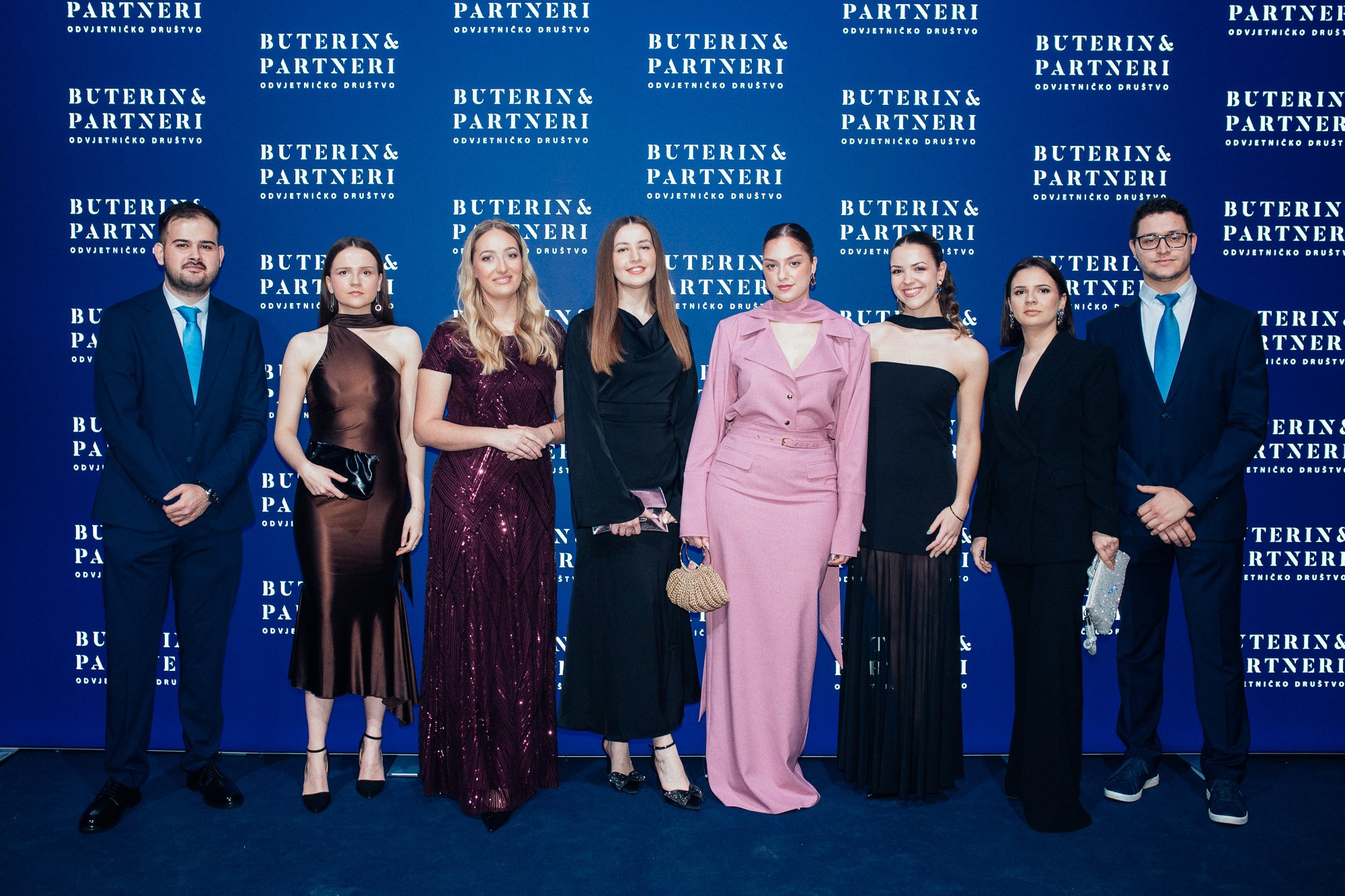 Group of nine young adults dressed in formal attire, standing in front of a blue backdrop with the logo and name 'Buterin & Partneri' repeated multiple times.