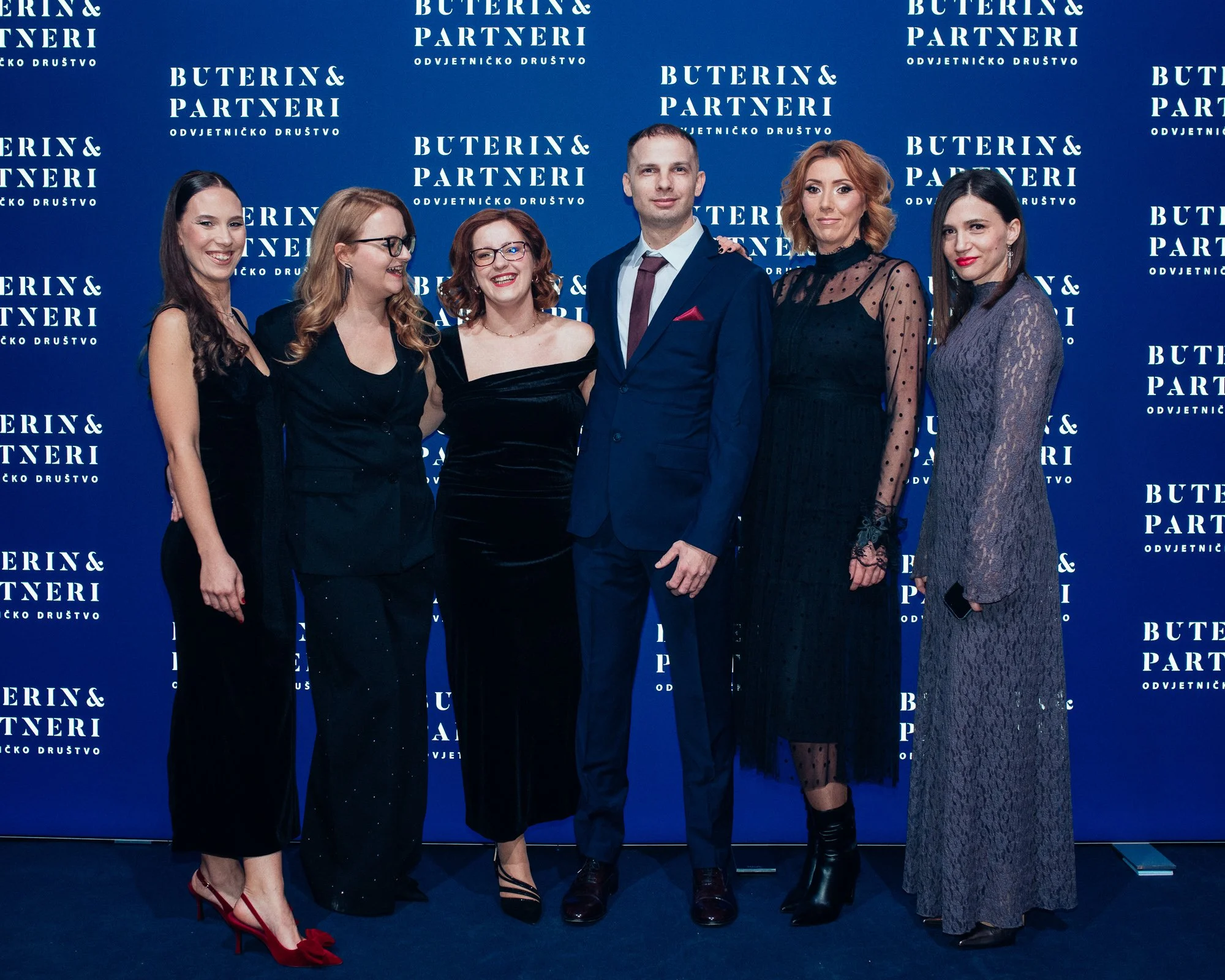 Six people standing together against a blue backdrop with white text that reads 'Buterin & Partneri Odvjetničko Društvo'. The group consists of five women and one man dressed in formal attire, smiling and posing for a photo at an event.