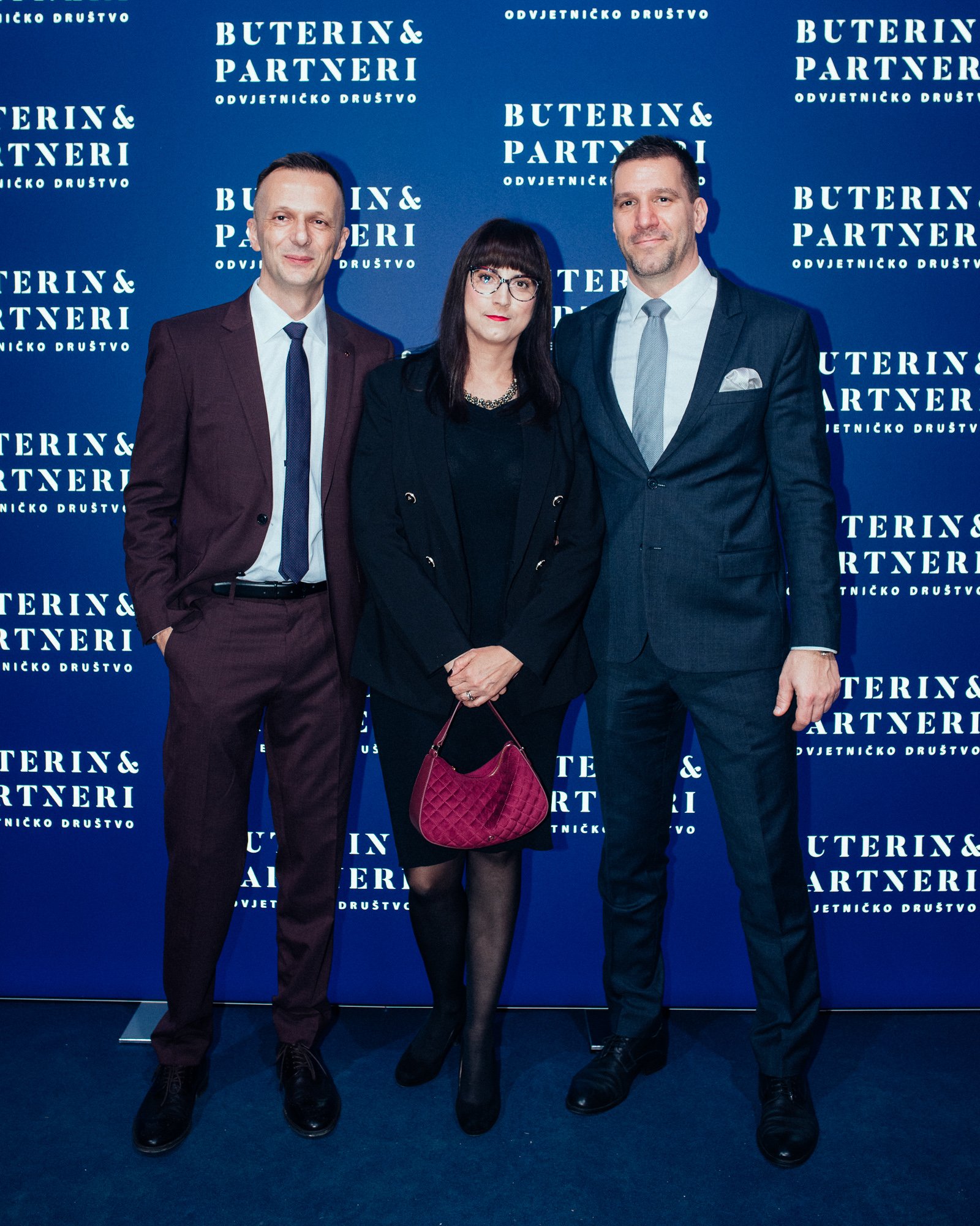 Three people standing in front of a blue backdrop with white text that reads "Buterin & Partneri Odvjetničko Društvo." The individuals are dressed in formal business attire, with two men wearing suits and a woman dressed in black holding a red quilte