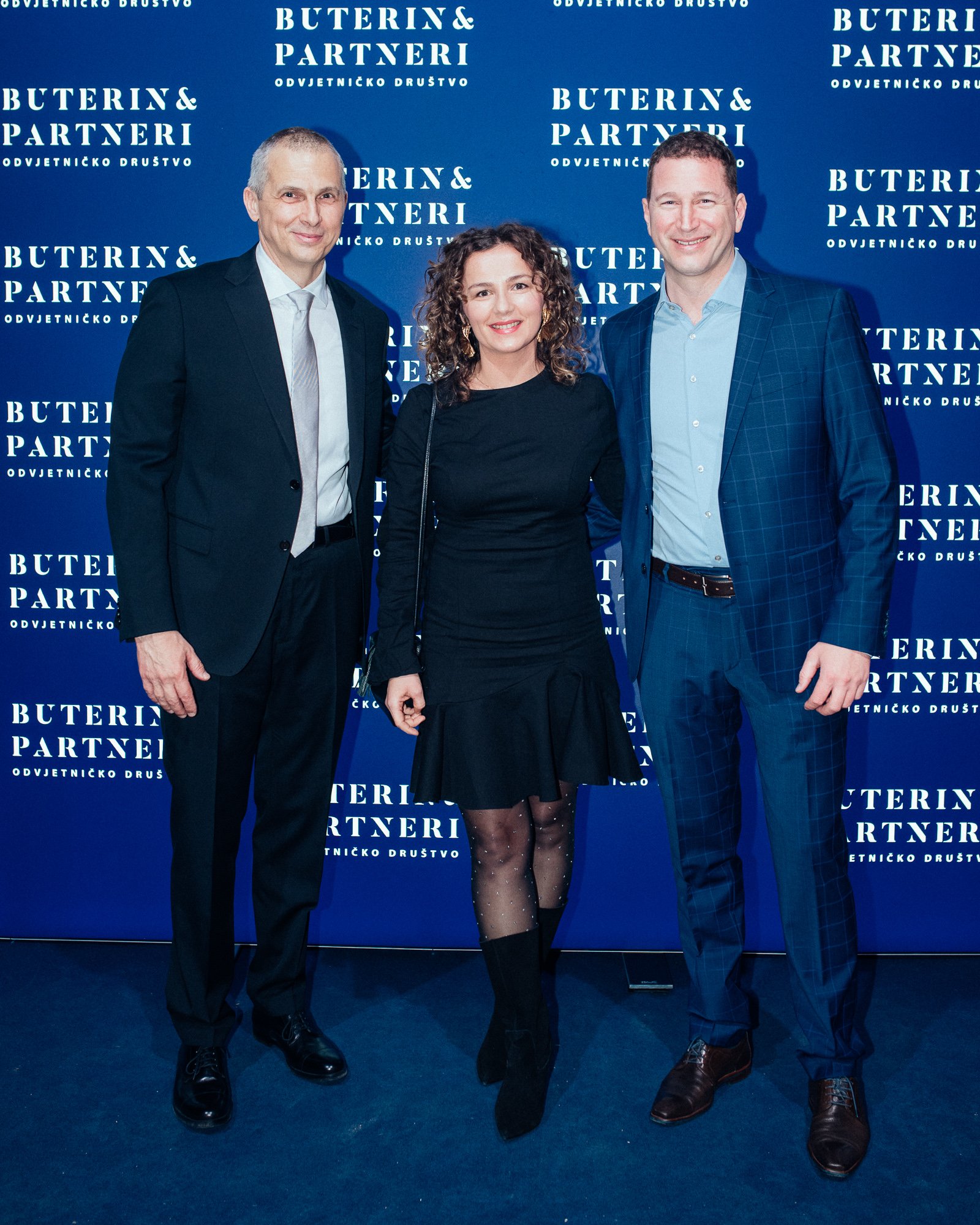 Three people standing together at a formal event in front of a blue backdrop with white text that says 'Buterin & Partneri' and 'Odvjetničko Društvo'. All are dressed in formal attire, smiling for the camera.