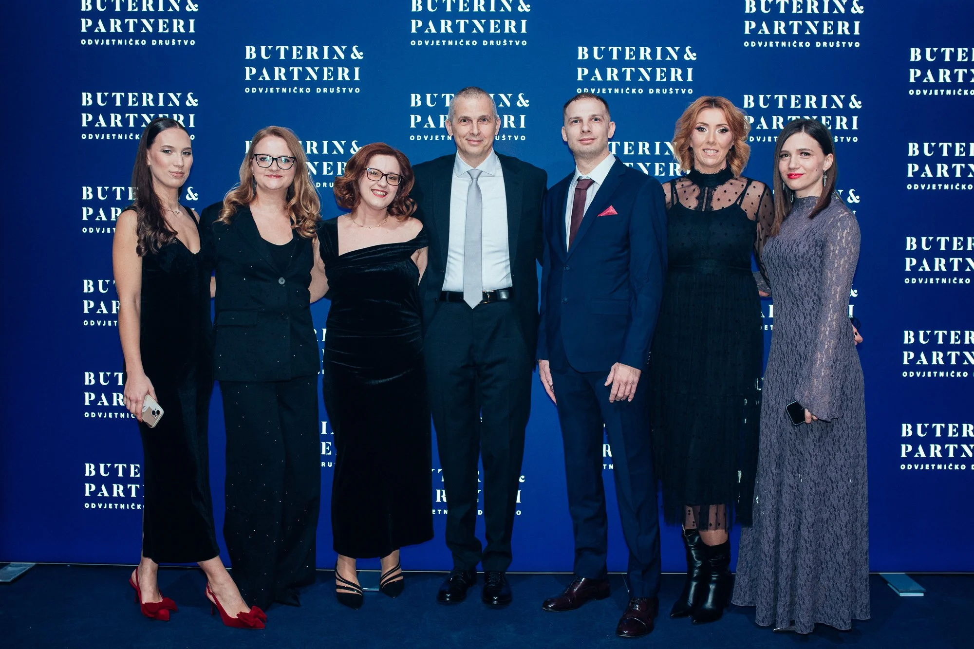 Group of seven well-dressed individuals at an event, standing in front of a blue backdrop with white text that reads 'BUTERIN & PARTNERI ODVJETNICKO DRUSTVO'.