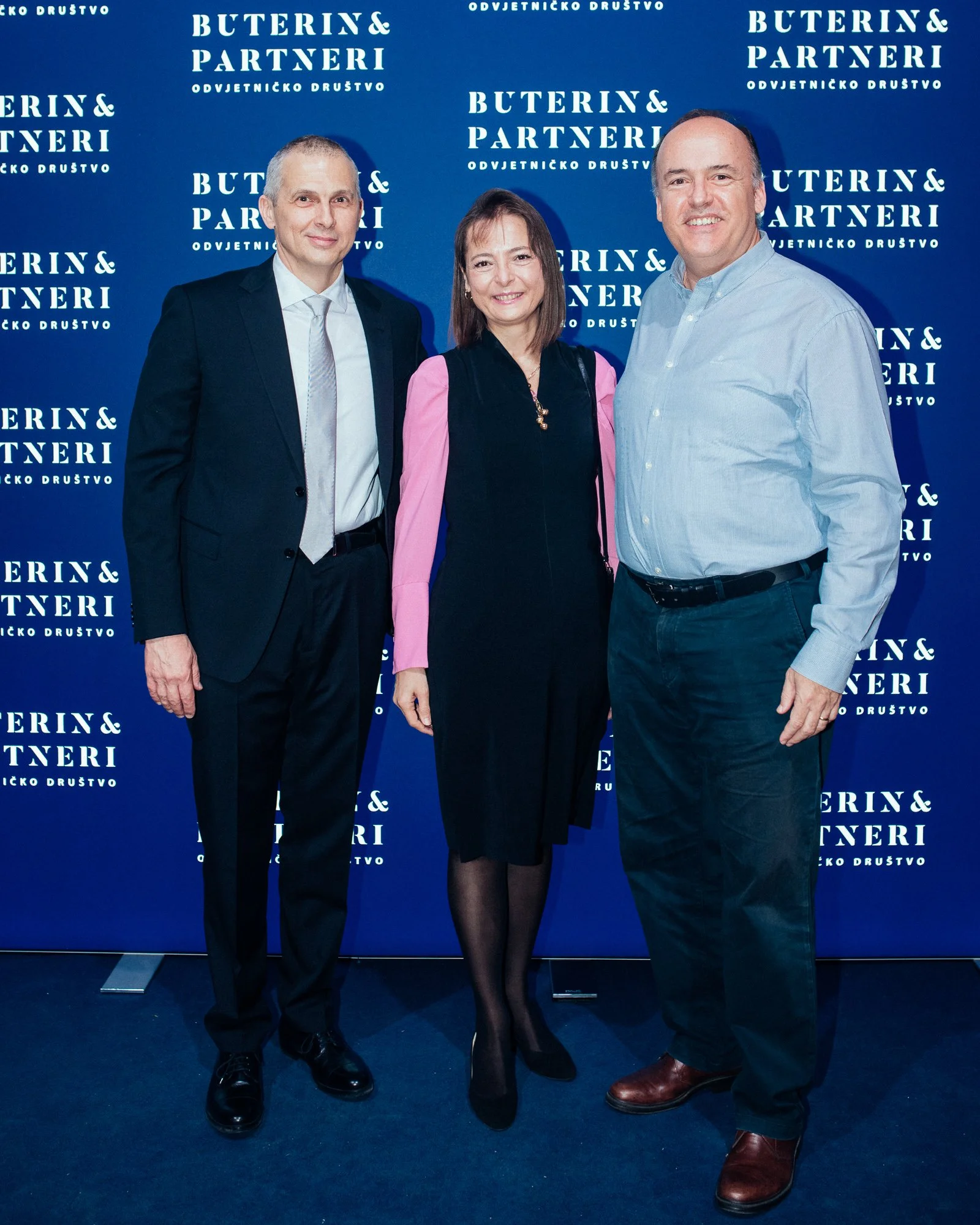 Three people standing together in front of a blue backdrop with white text, at an event. The man on the left is wearing a black suit with a light blue shirt and tie. The woman in the center is wearing a black dress with pink sleeves and black tights.