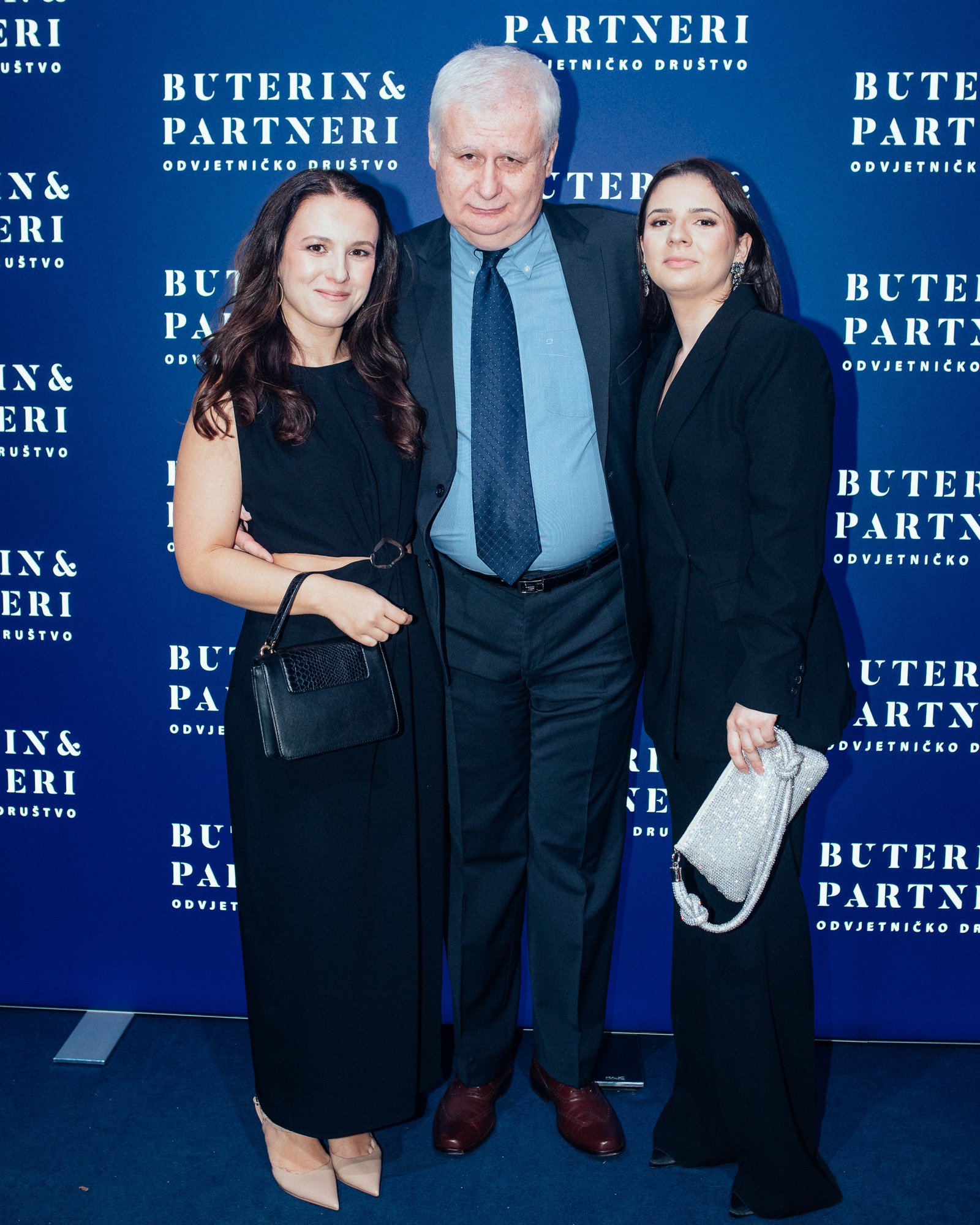 Three people standing in front of a blue backdrop with white text that reads 'Buterin & Partneri' and other words. The person in the middle is an older man with white hair, wearing a dark suit, light blue shirt, and blue tie. The woman on the left ha