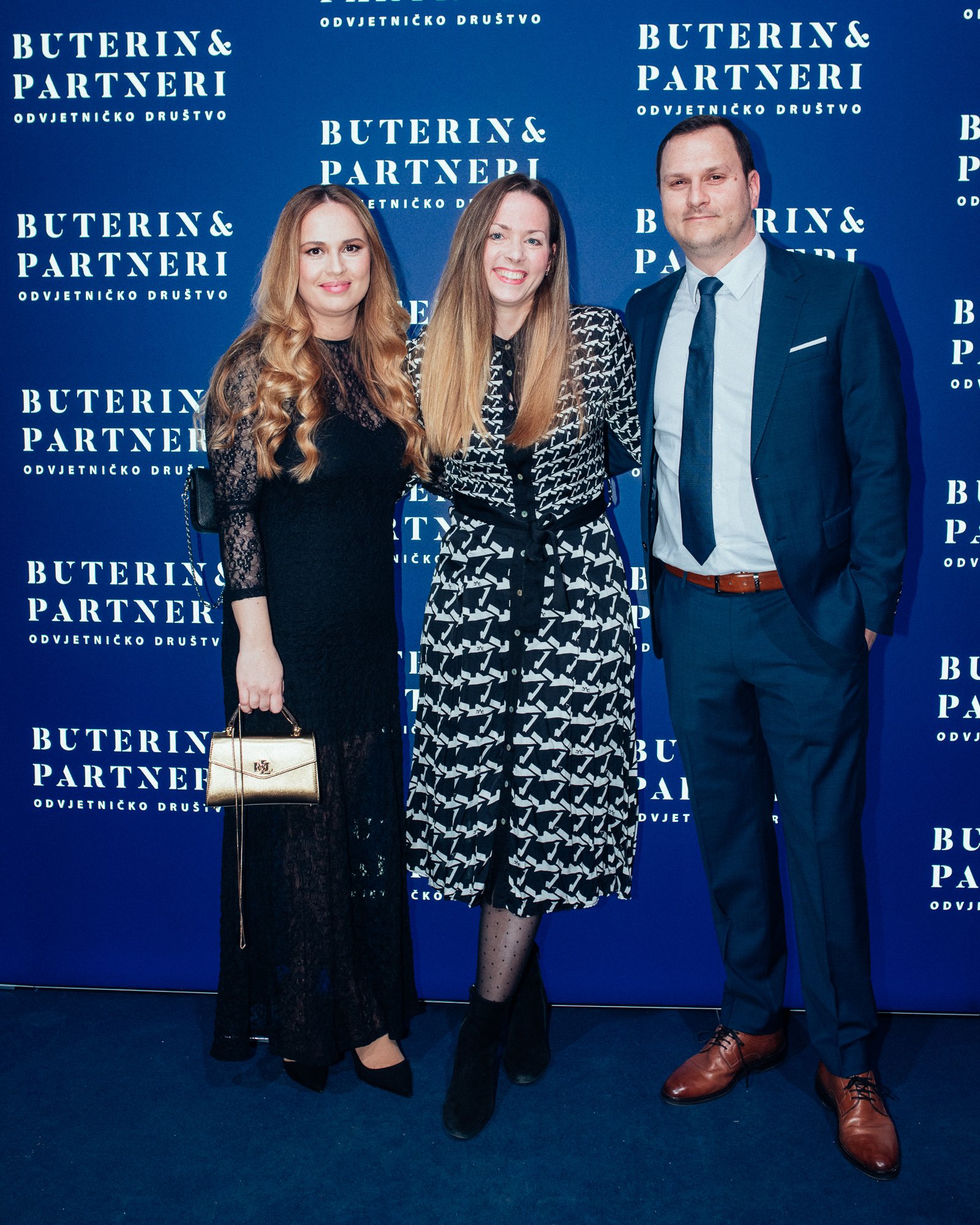 Three people standing in front of a blue backdrop with white text that reads 'Buterin & Partneri' and 'Odvjetničko Društvo.' The two women are wearing black dresses, and the man is in a blue suit with a light blue shirt and dark tie.