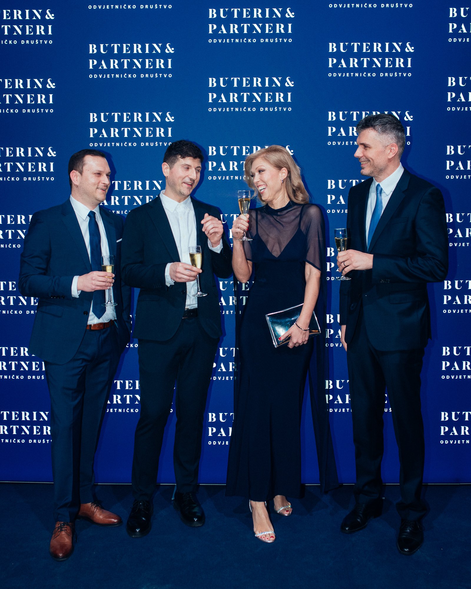 Four people in formal attire standing in front of a blue backdrop with the text 'BUTERIN & PARTNERI' and 'ODVJETNIČKO DRUŠTVO'. They are holding glasses of champagne and appear to be at a celebration or event, engaging in conversation and smiling.