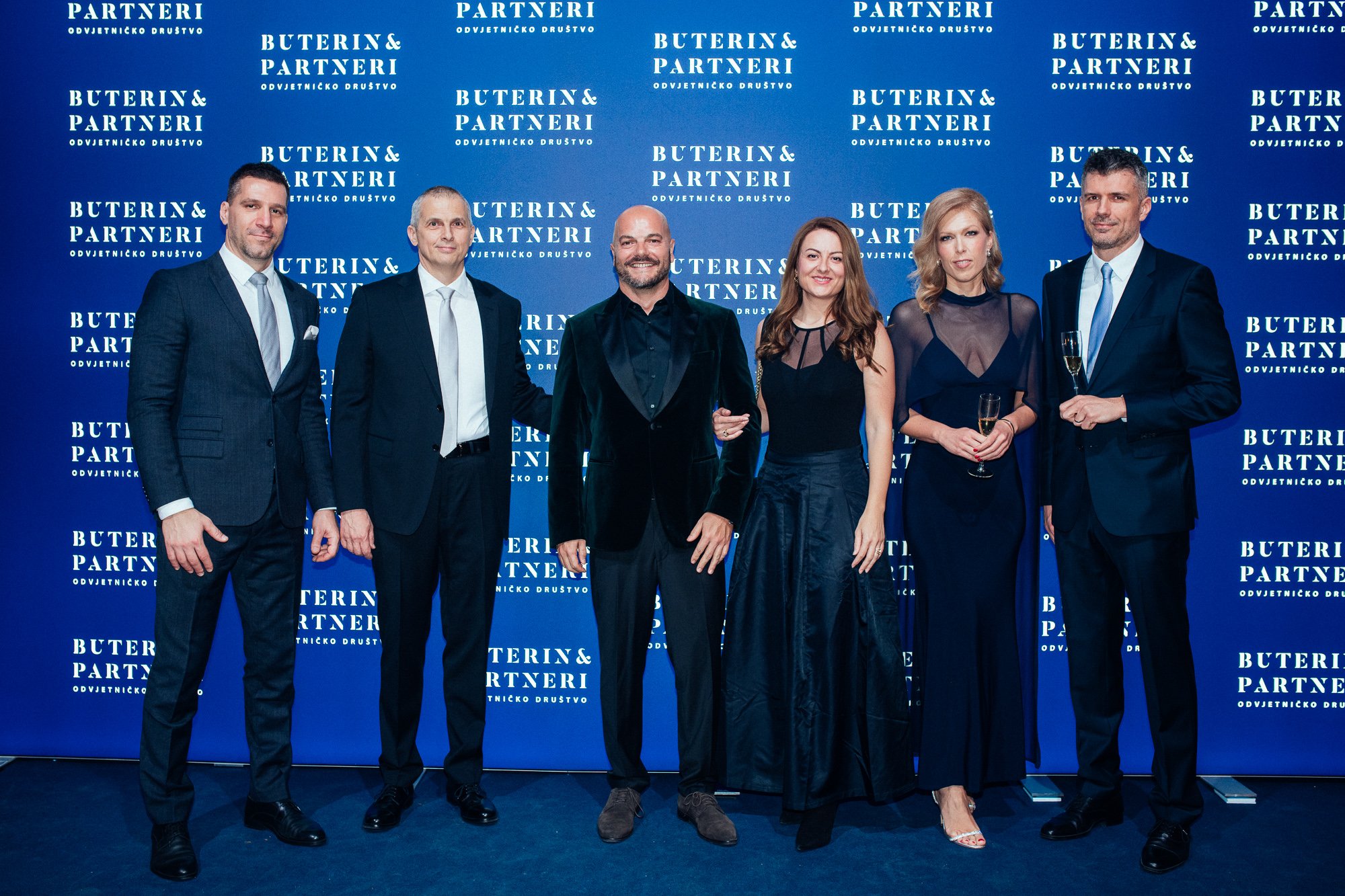 Group of six people dressed in formal attire standing in front of a blue backdrop with the text "Buterin & Partneri" repeated multiple times, likely at a formal event or celebration.