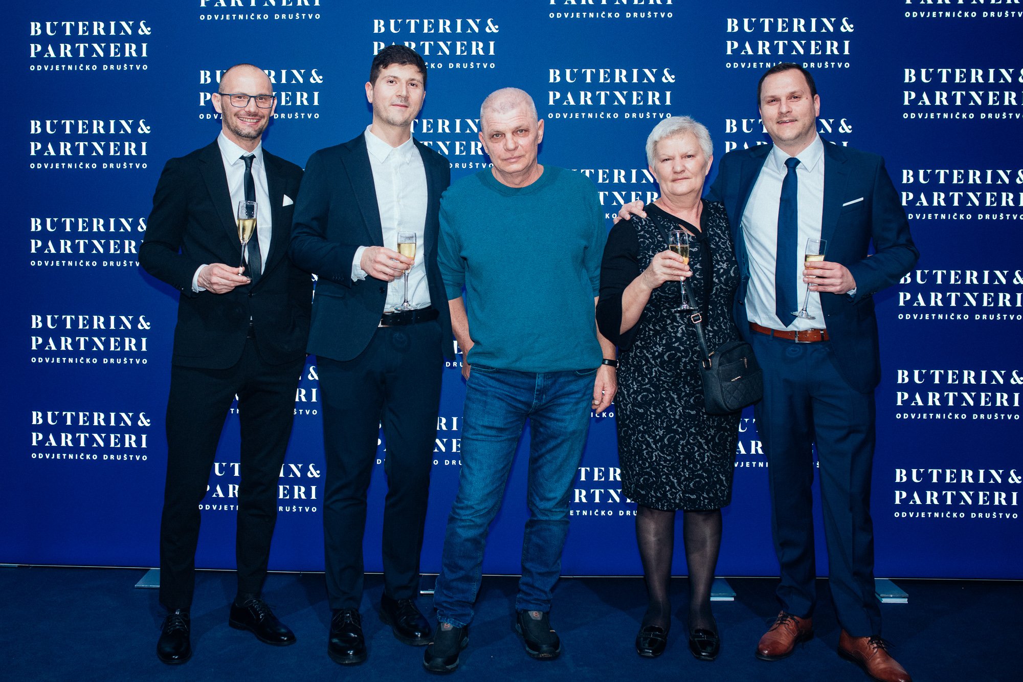 Six people standing in front of a blue backdrop with white text that reads "Buterin & Partneri" and "Odvjetničko društvo." The group is dressed in formal attire, holding glasses of champagne, at an event or celebration.
