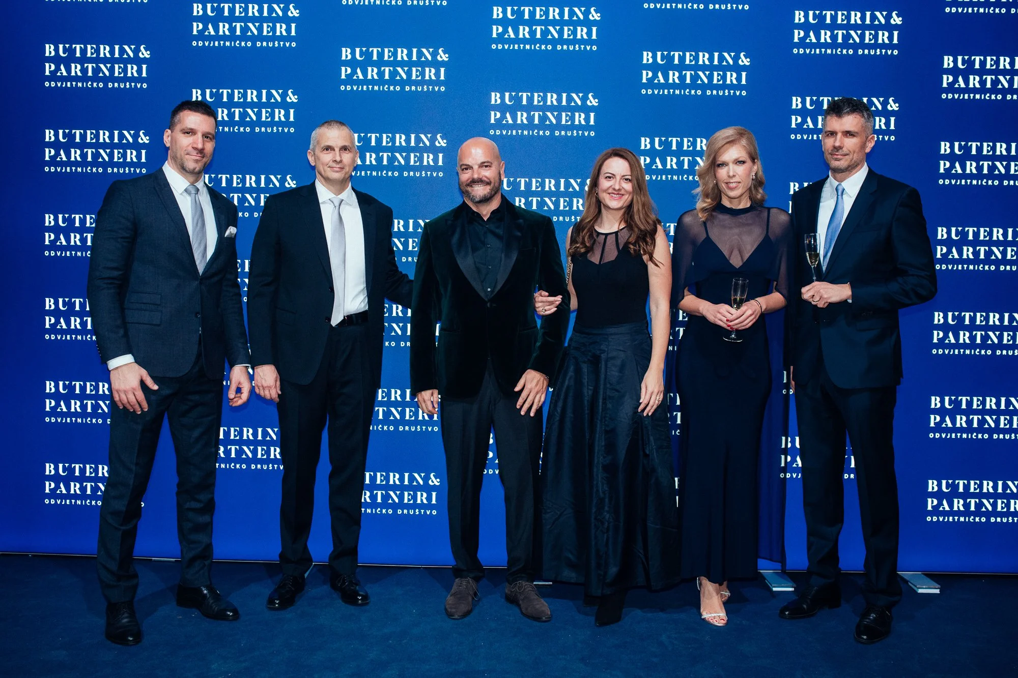 Six people in formal attire standing in front of a blue backdrop with the text "Buterin & Partneri" repeated in white. They are smiling, and two of them are holding champagne glasses.