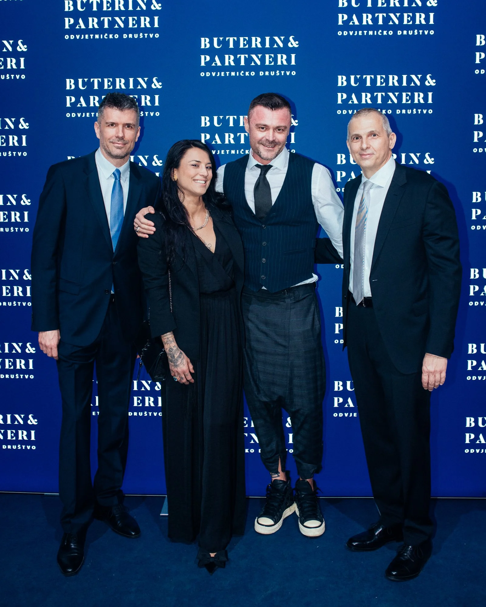 Four people standing in front of a blue backdrop with white text that reads 'Buterin & Partneri Odvjetničko Društvo', posing for a photo at a formal event. The group includes three men in suits and a woman in black attire.