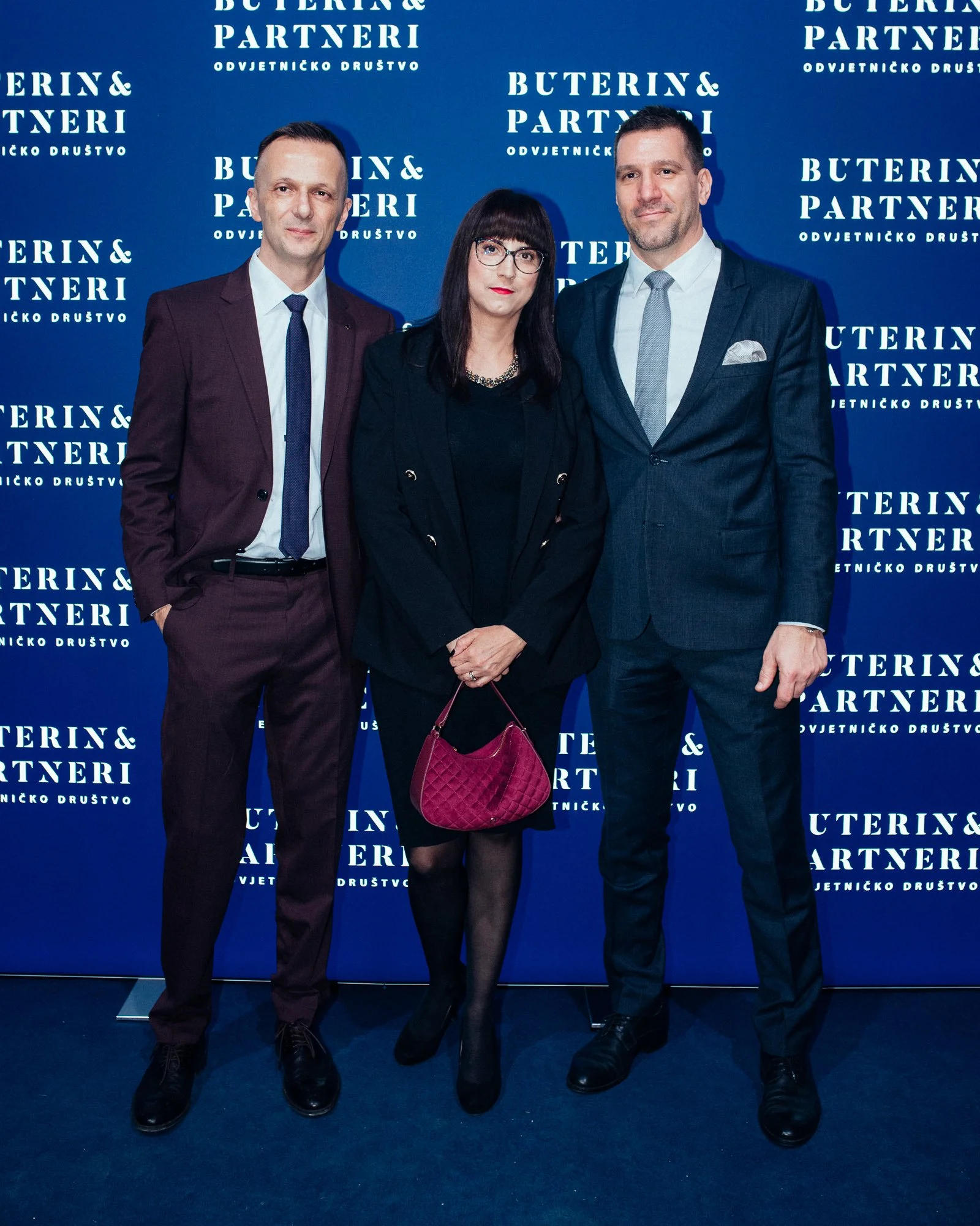 Three professionally dressed individuals standing in front of a blue backdrop with white text repeating 'Buterin & Partneri Odvjetničko Društvo.' The man on the left is wearing a dark maroon suit with a white shirt and tie, the woman in the middle is