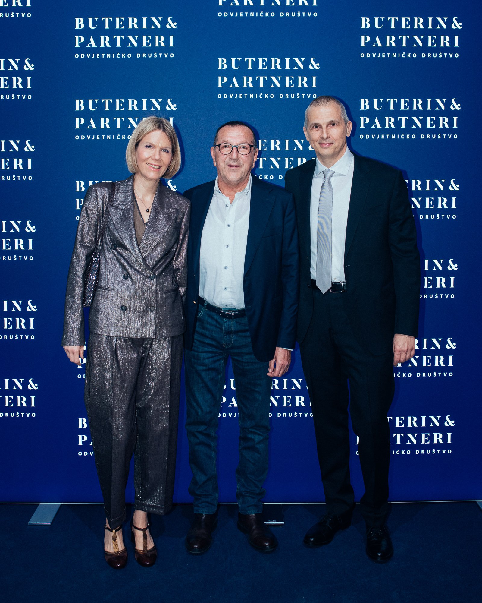 Three people, two men and one woman, standing together in front of a blue backdrop with white text that reads 'Buterin & Partneri Odvjetničko društvo.'