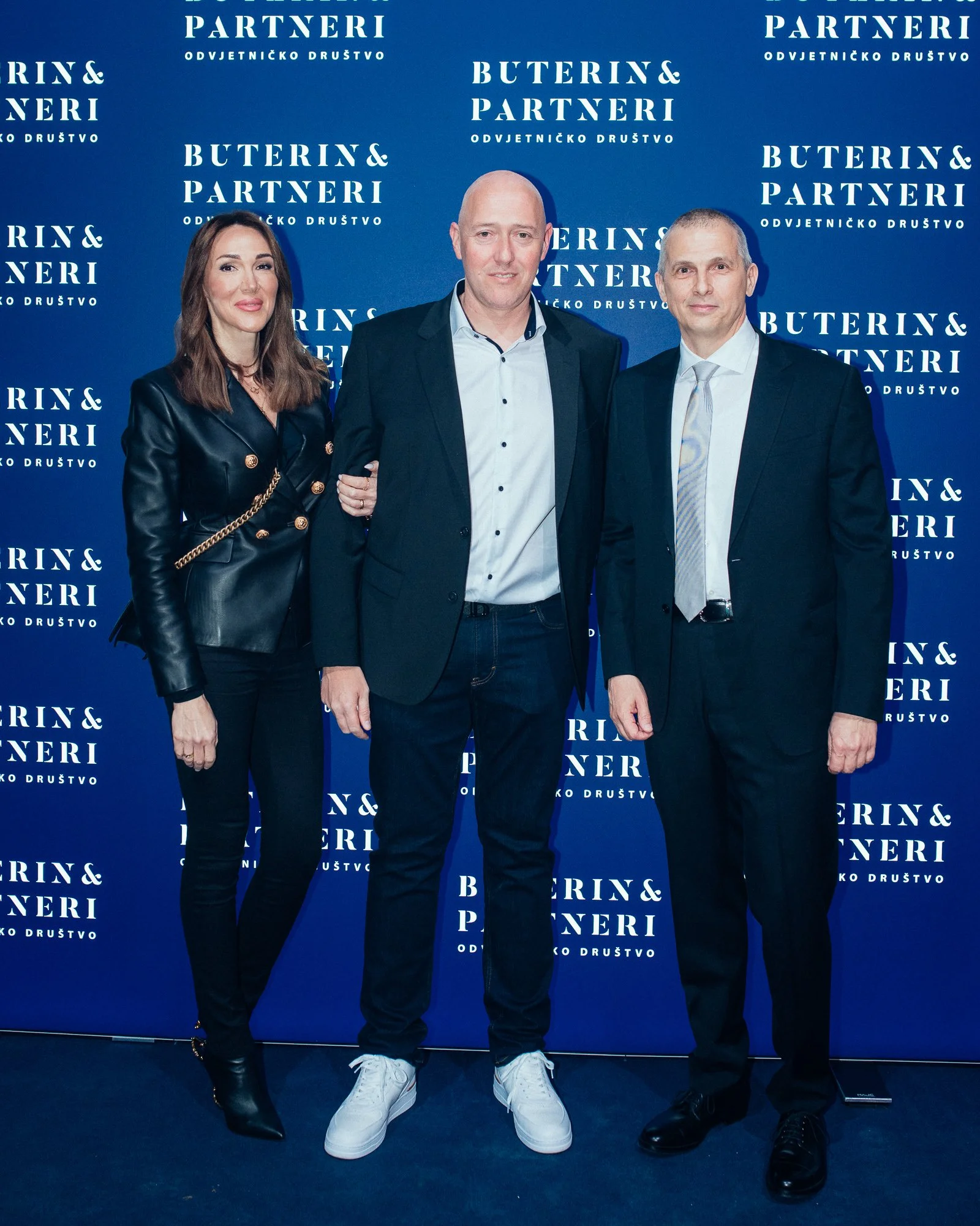 Three people standing in front of a blue backdrop with white text that says "Buterin & Partneri" and "Odvjetničko Društvo." The woman on the left has long brown hair and is wearing a black leather blazer with gold buttons and black pants. The man in 
